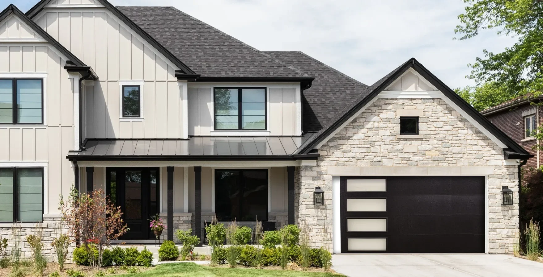 Modern two-story house with white siding, stone accents, and a black garage door with frosted glass panels, surrounded by a landscaped garden.