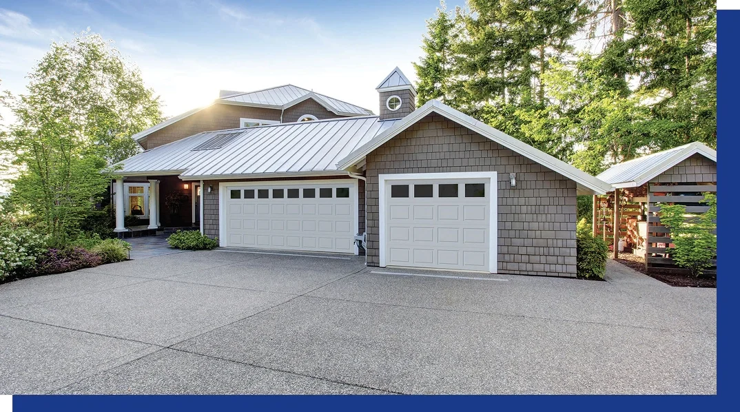 Front view of a modern house with a concrete driveway, gray siding, white garage doors, and surrounded by trees and greenery.