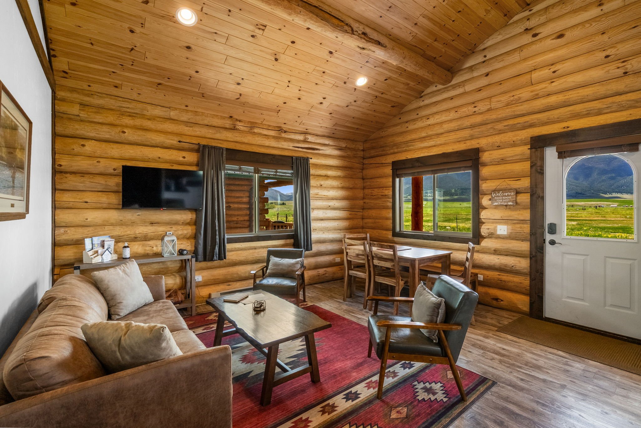 A cozy bedroom featuring a bed with white bedding, gray blanket, and pillows, wooden walls, window with blinds and dark curtains, potted plant, nightstand with a lamp, and framed artwork on the wall. Near Yellowstone, Chico, Old Saloon, and great fis