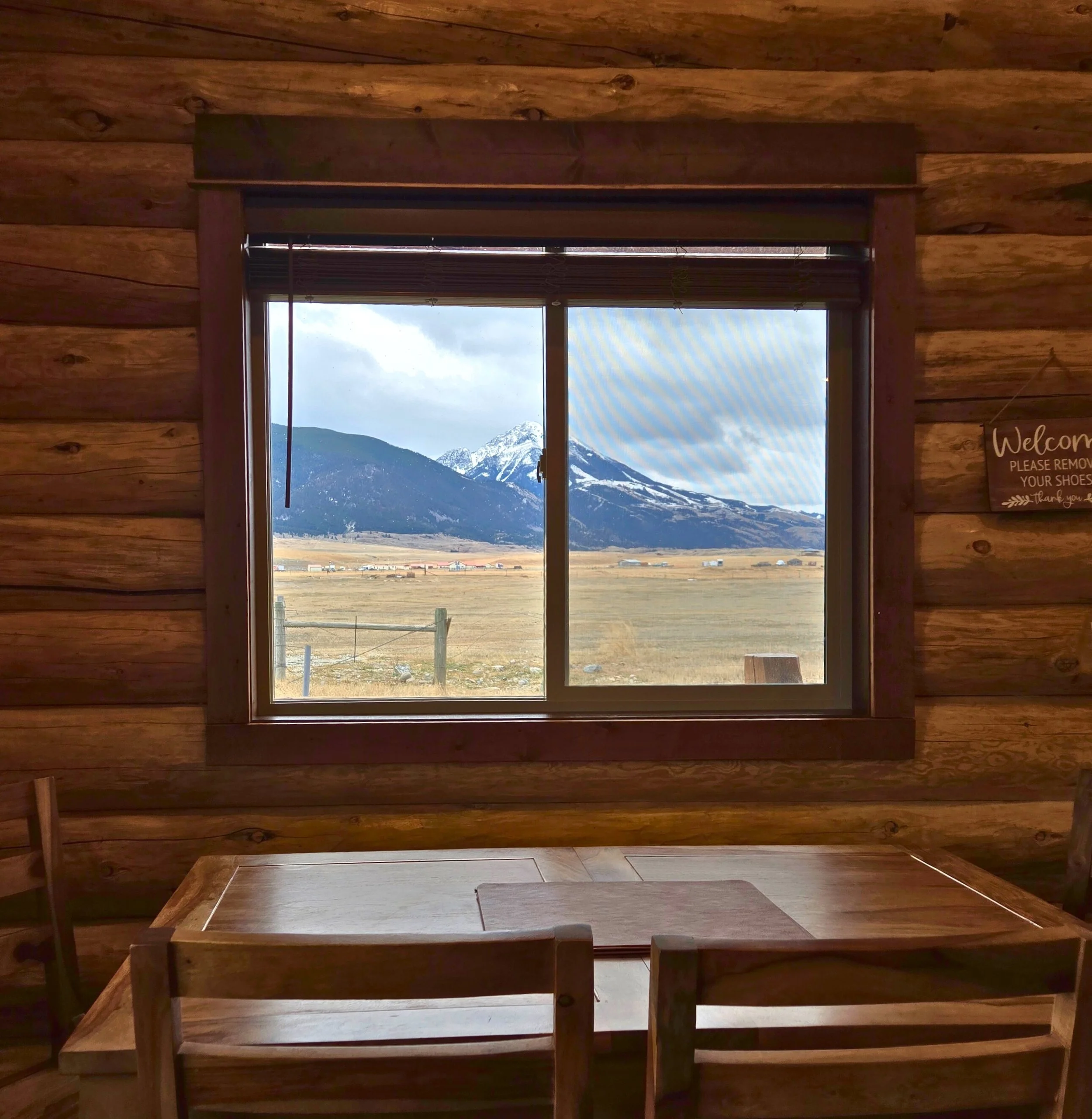 Dining area with a wooden table set with black dishes, silverware, and glasses, a vase with green and red foliage, in front of a large window with blinds showing mountains and blue sky outside. Near Yellowstone, Chico, Old Saloon, and great fishing.