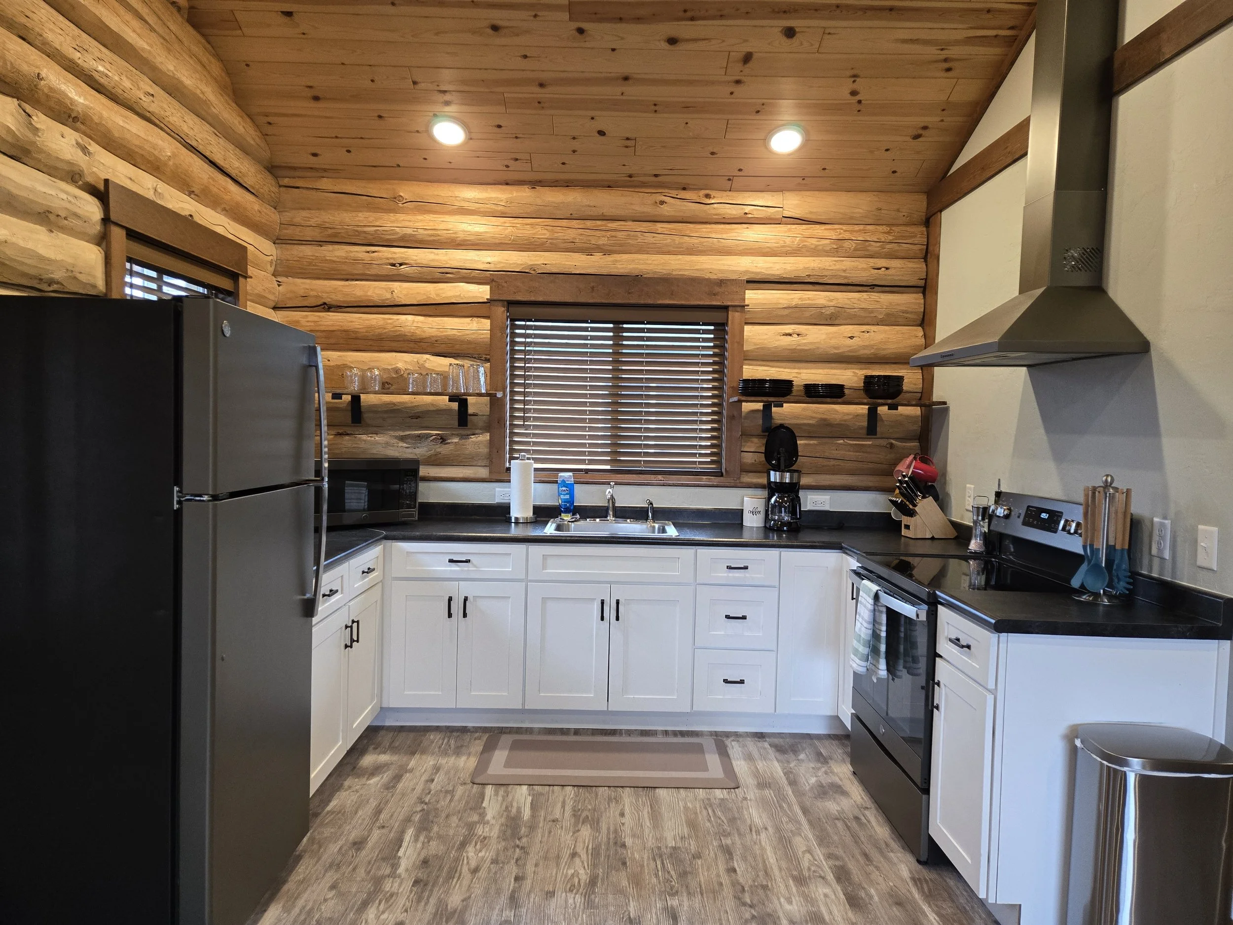 A cozy bedroom featuring a bed with white bedding, gray blanket, and pillows, wooden walls, window with blinds and dark curtains, potted plant, nightstand with a lamp, and framed artwork on the wall. Near Yellowstone, Chico, Old Saloon, and great fis
