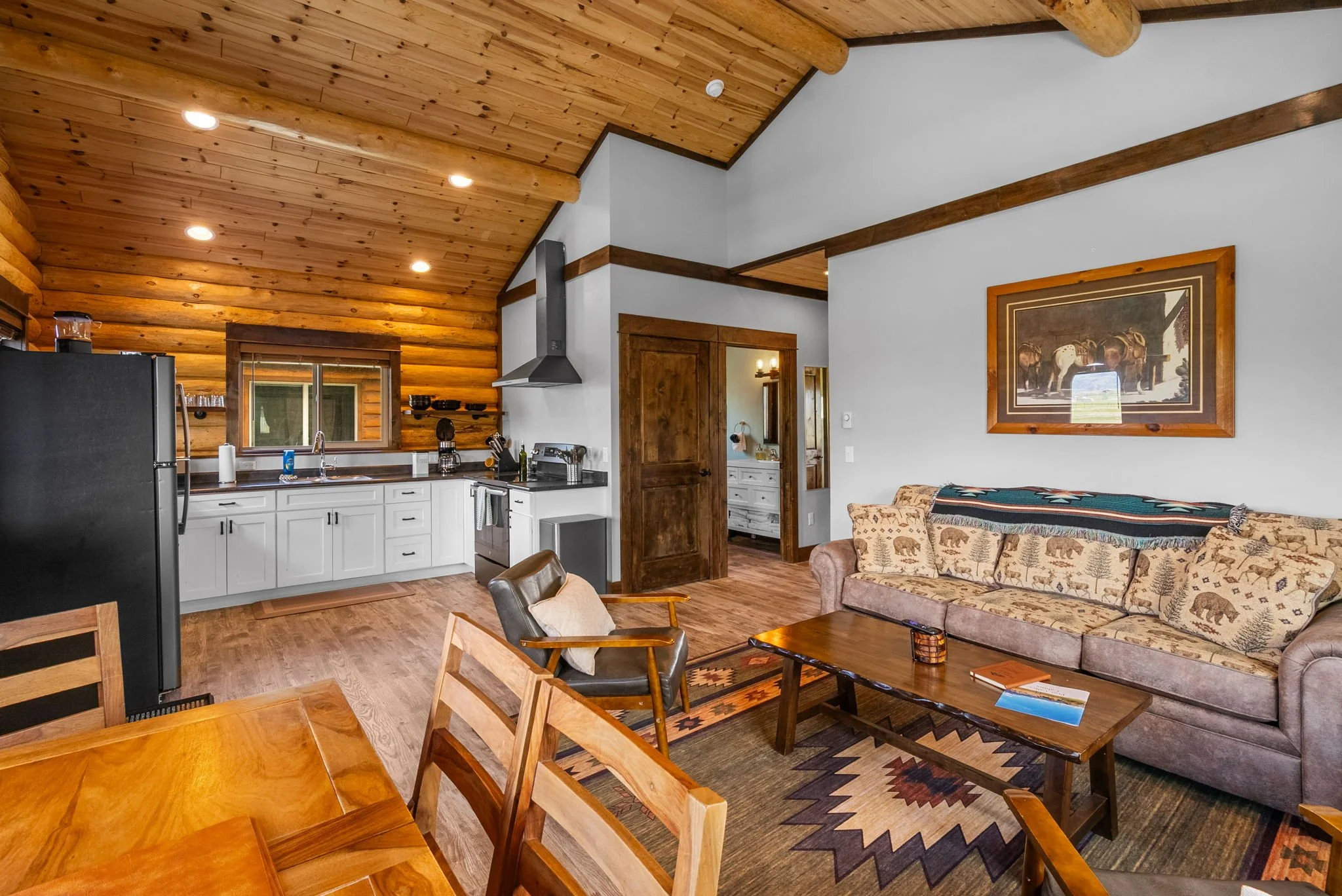 Dining area with a wooden table set with black dishes, silverware, and glasses, a vase with green and red foliage, in front of a large window with blinds showing mountains and blue sky outside. Near Yellowstone, Chico, Old Saloon, and great fishing.