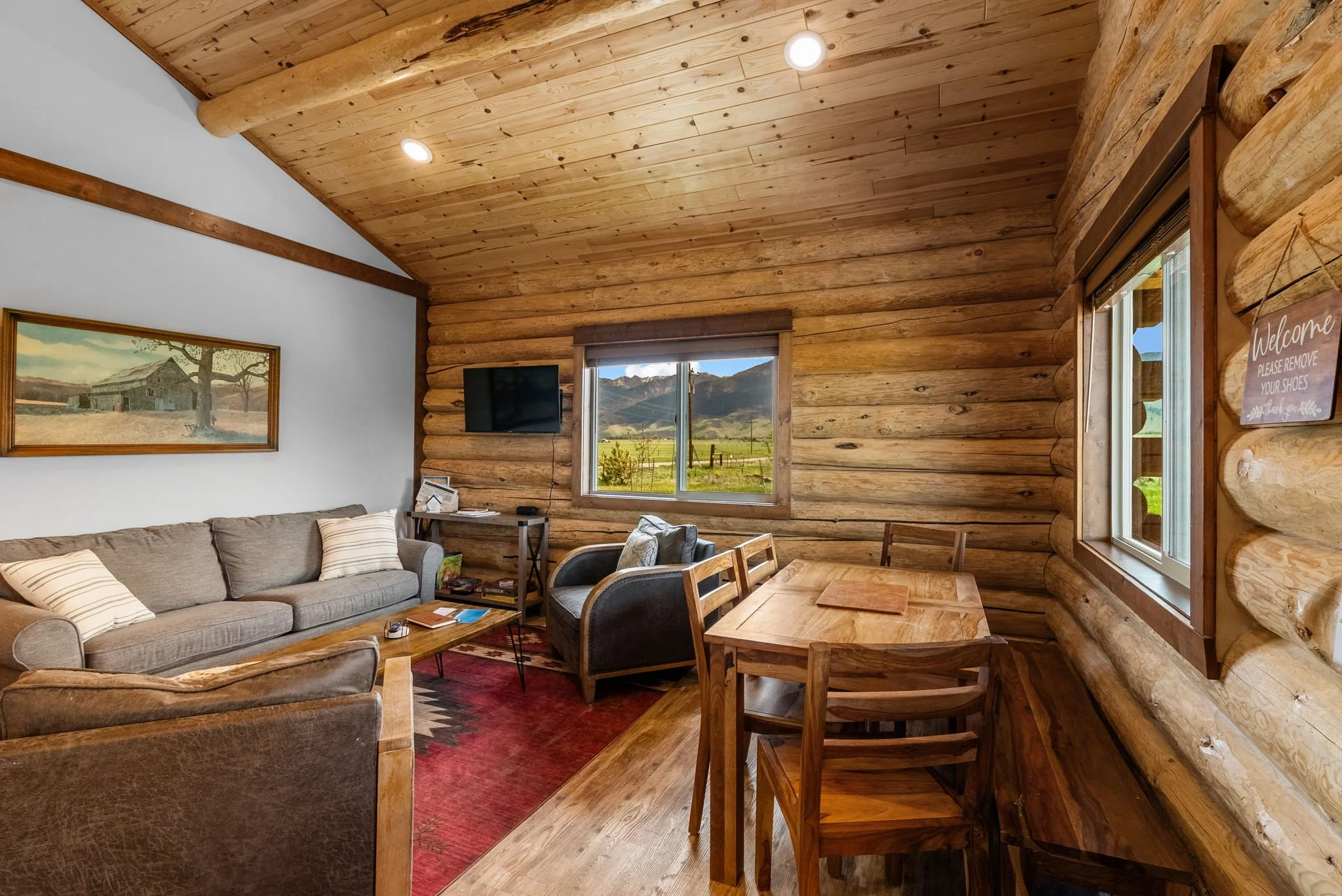 A cozy bedroom with wooden walls, a bed with white bedding, a nightstand with a lamp, a window with blinds, and a colorful framed picture on the wall. Near Yellowstone, Chico, Old Saloon, and great fishing.