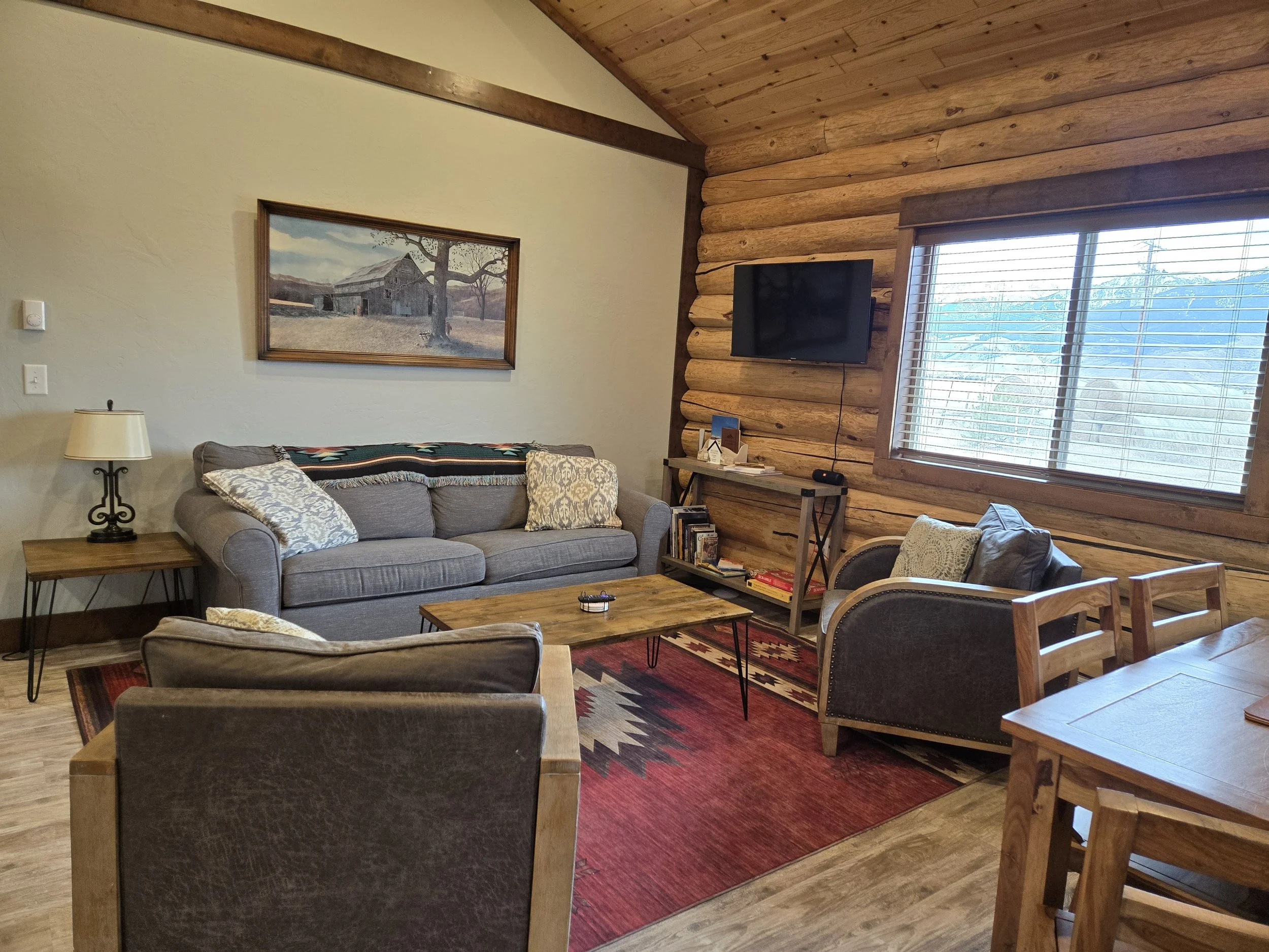 A cozy bedroom with wooden walls, a bed with white bedding, a nightstand with a lamp, a window with blinds, and a colorful framed picture on the wall. Near Yellowstone, Chico, Old Saloon, and great fishing.