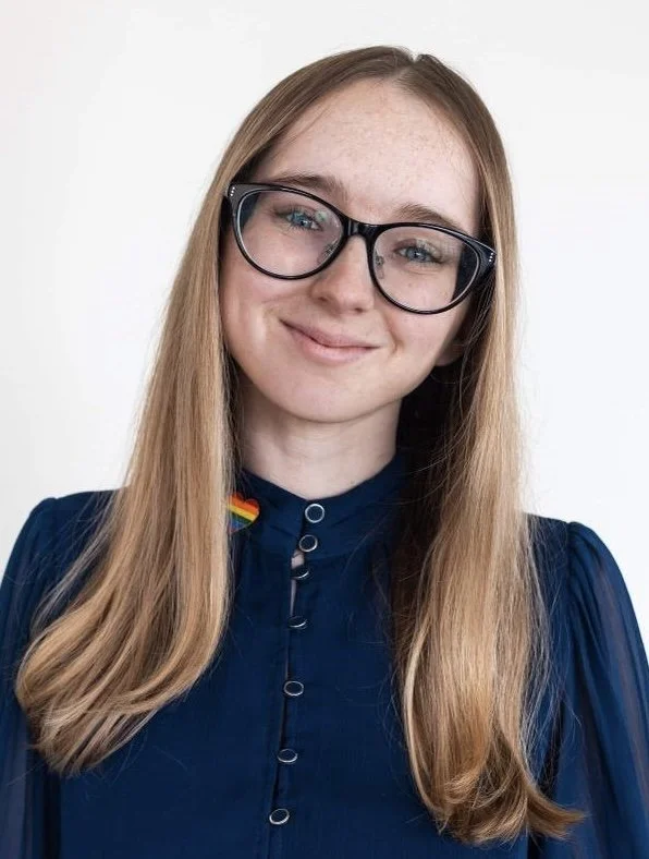 Young woman with long blonde hair wearing glasses and a dark blue top with a rainbow pride ribbon pin, smiling at the camera.