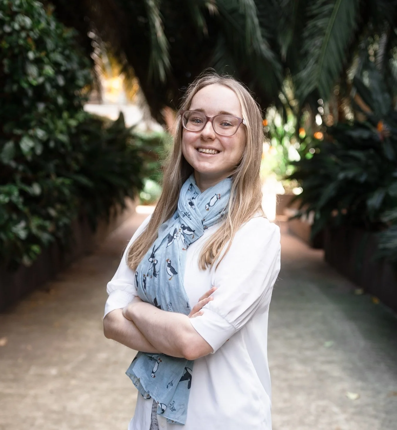 Experienced Counsellor: Liz O'Neill.  A young woman with glasses and long blonde hair smiling with arms crossed, standing in a lush garden pathway with a blue puffin scarf to showcase her Among Puffins Counselling Business.