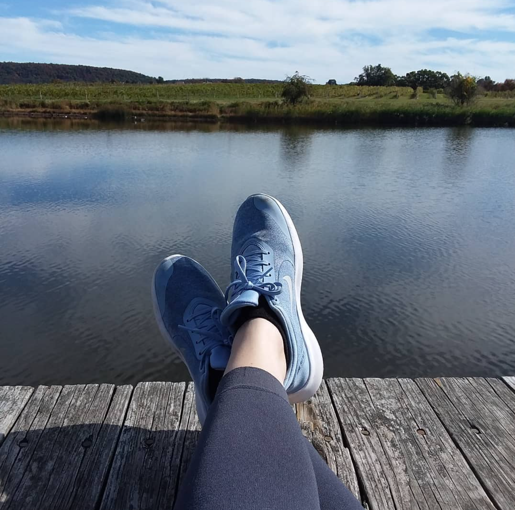Person relaxing on a wooden dock by a calm river, wearing blue sneakers and gray leggings, with a scenic landscape of trees and rolling hills in the background under a partly cloudy sky.