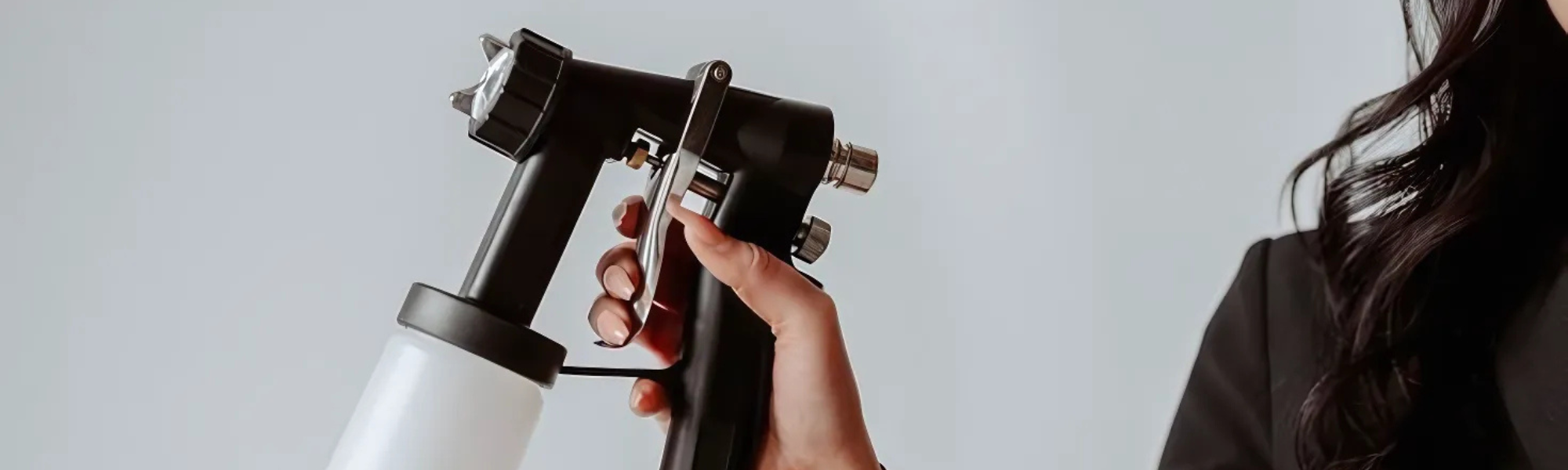 A brunette woman holding a spray tan machine applicator. She is standing in front of a white background, wearing a black blazer.