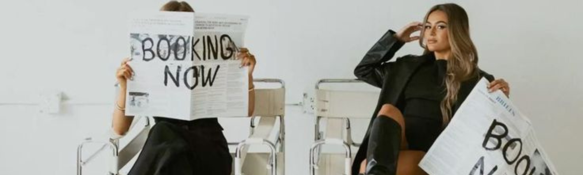 Two women sitting in white metal chairs, holding newspapers that say "book now" they are posing for a brand photoshoot to attract clients to their beauty business