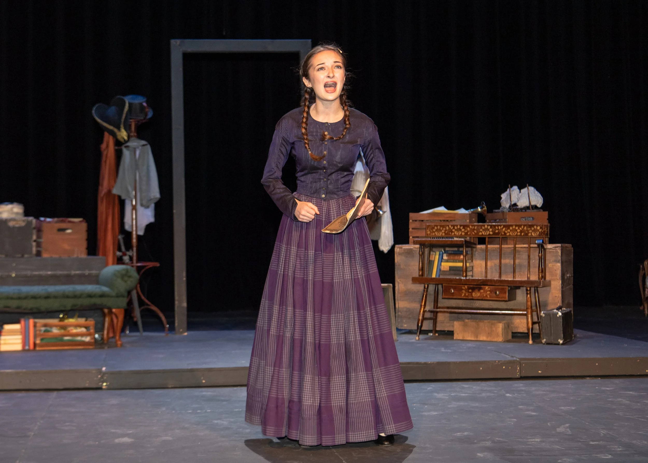 Sarah Novak in Little Women at Kent Denver School. Woman in period costume-like dress singing on stage with vintage furniture and props in background.