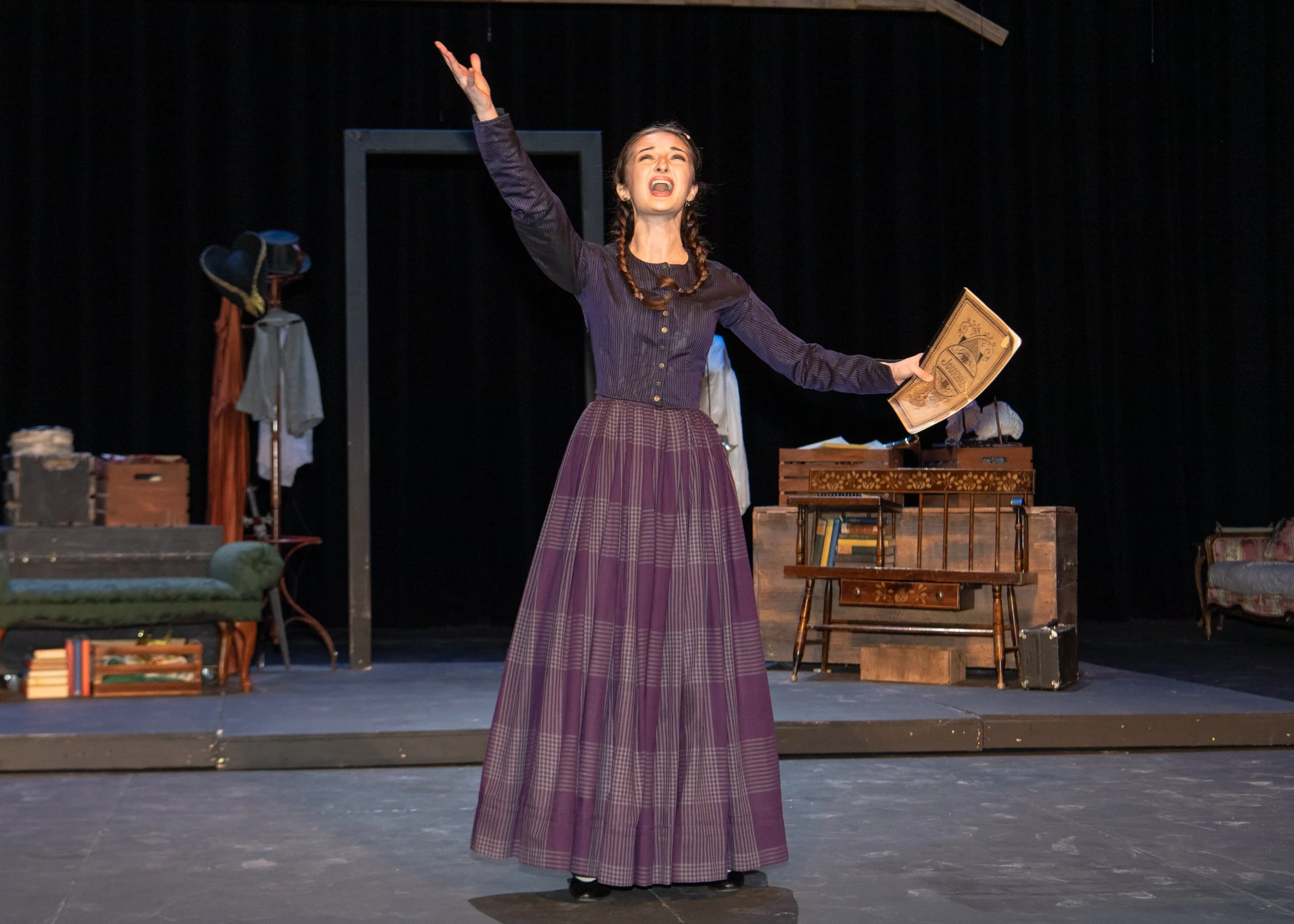 A woman dressed in period costume on stage with her arm raised, holding a newspaper, gesturing passionately, in a theatrical performance with vintage furniture and props around her.