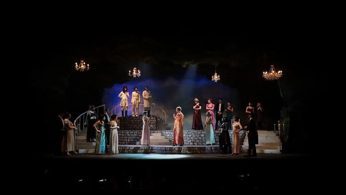 The image shows a theatrical stage set resembling an outdoor garden with a stone staircase, where multiple performers are dressed in vintage formal attire, participating in a theatrical production. Chandeliers hang from above, enhancing the elegant ambiance, and stage lighting highlights the performers.