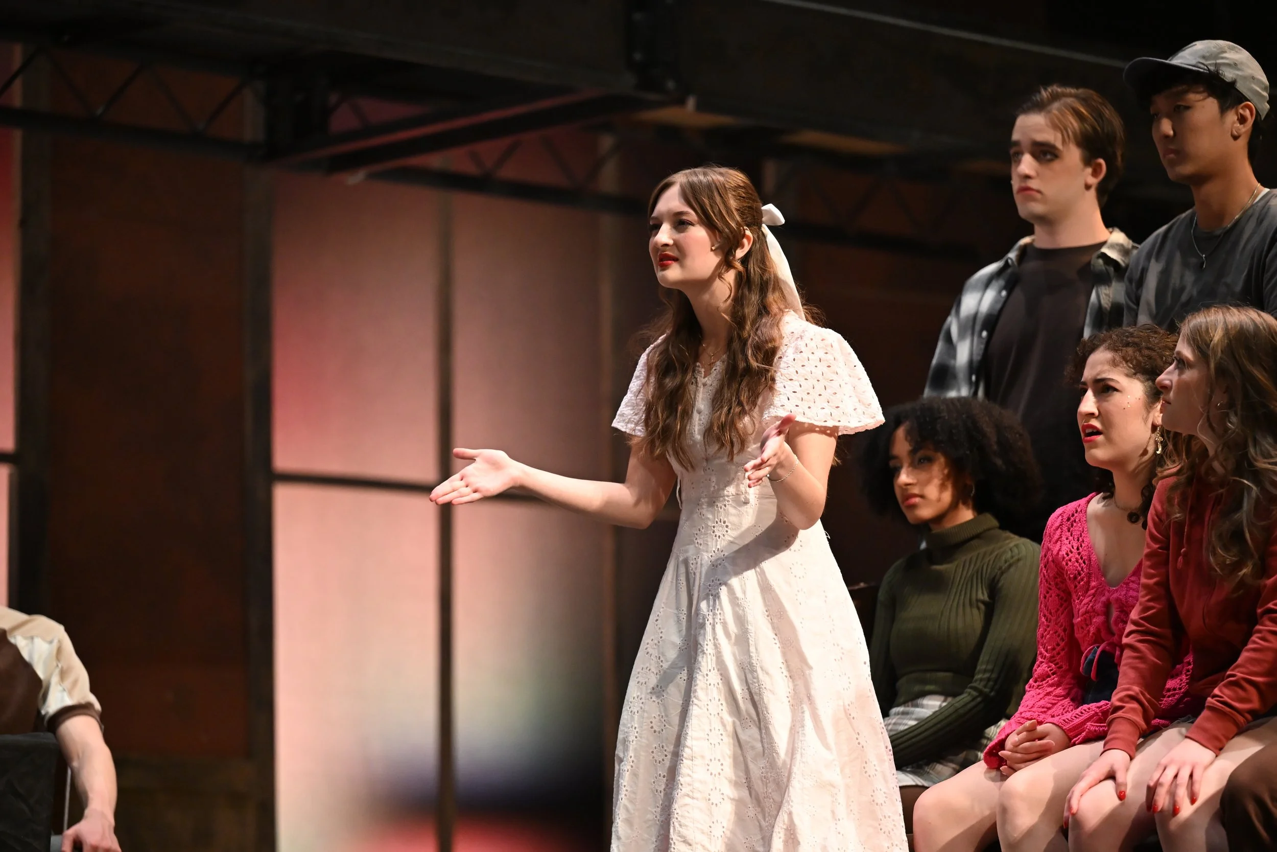 A group of young actors on stage during a rehearsal or performance, with Sarah Novak in a white dress speaking and gesturing, and others seated behind her, watching attentively.