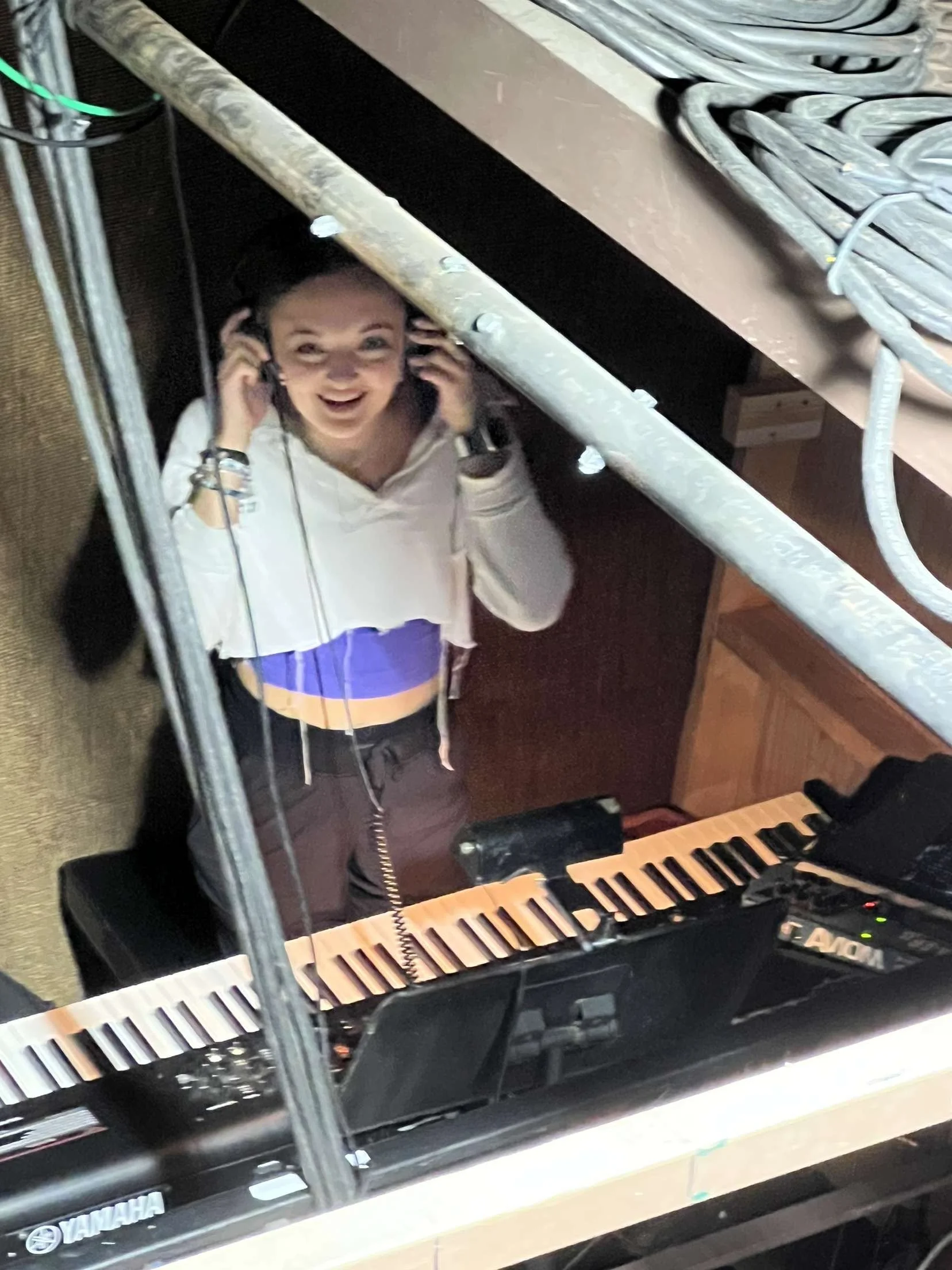 A young woman wearing headphones, a white jacket, and a colorful top, standing behind a keyboard synthesizer, smiling and looking up at the camera through a glass reflection.