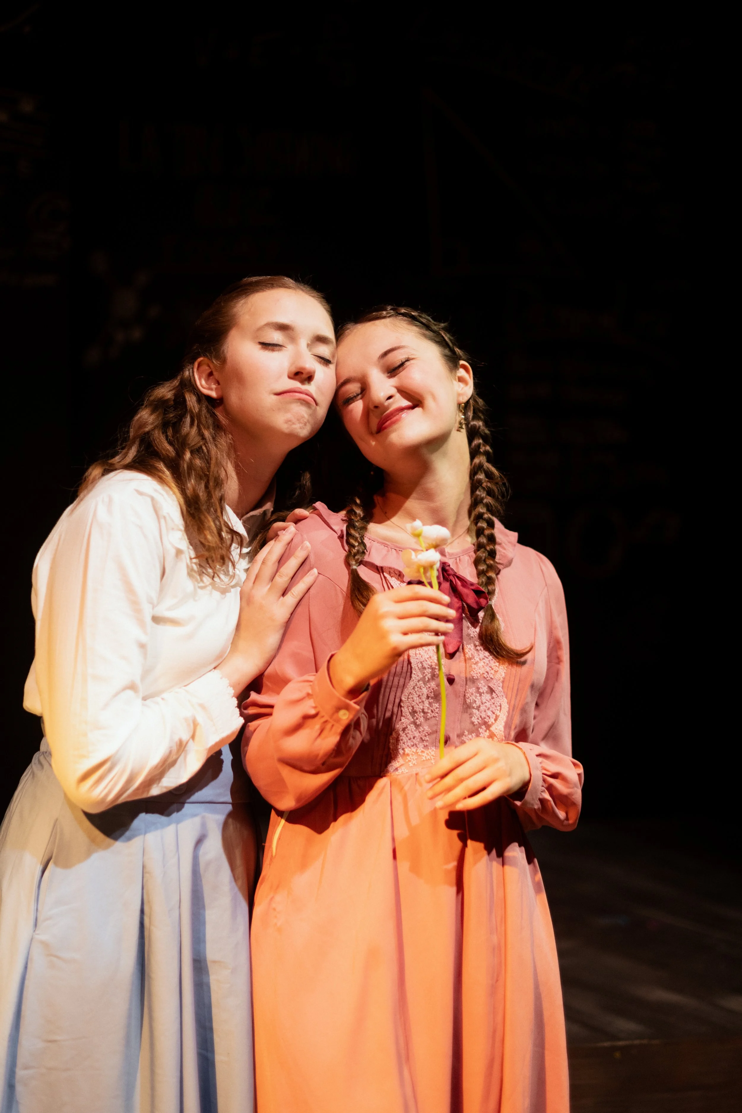 Sarah Novak in Spring Awakening at Point2Point Productions. Two young women with braided hair, wearing vintage dresses, standing close together on a stage, with one holding a flower and both smiling contentedly.