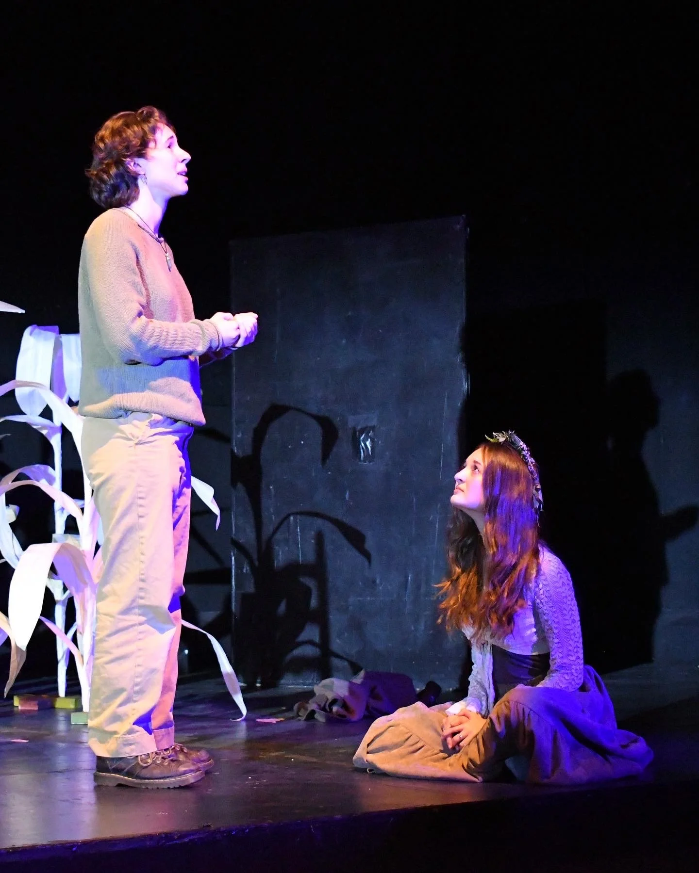 Two young women performing a theatrical scene on stage, one standing and speaking, the other kneeling and listening, with stage props and dark background.