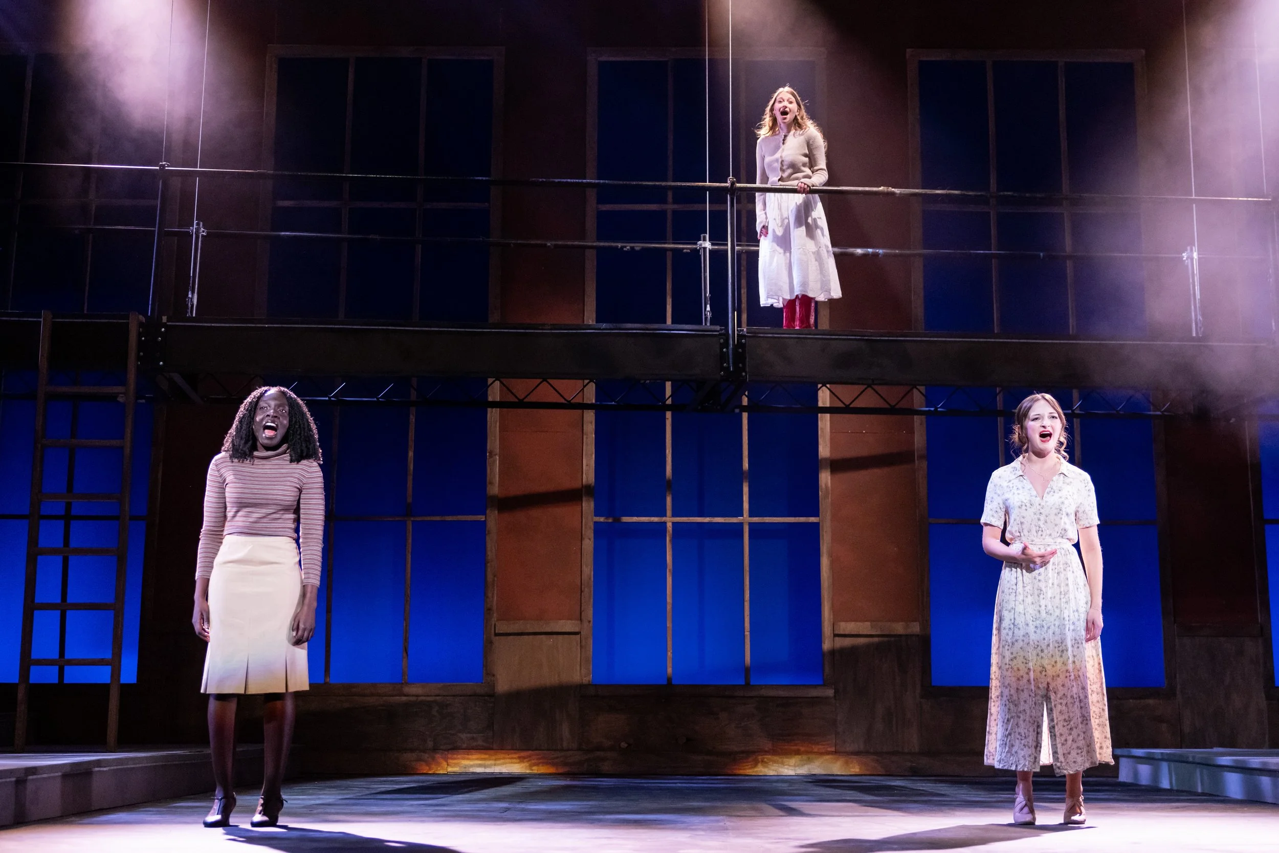 Three women performing on a theater stage, with one on the upper level and two on the lower level, all singing or speaking passionately.