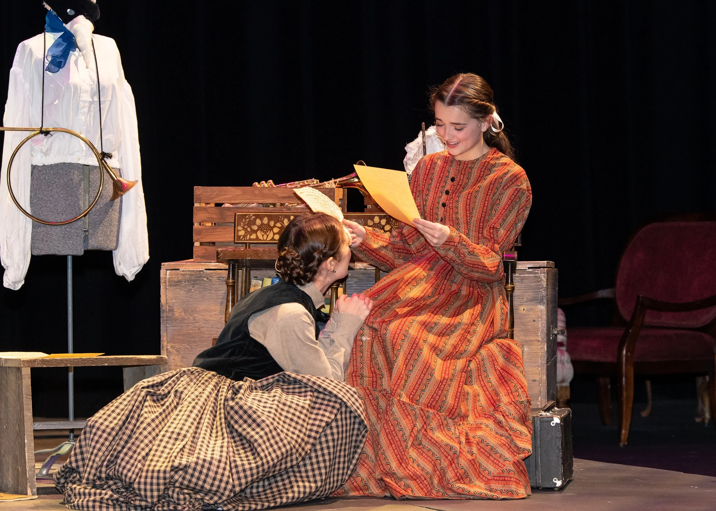 Two women in period costumes, one sitting on a wooden crate reading a paper and the other kneeling, in a theater scene with vintage furniture and clothing.