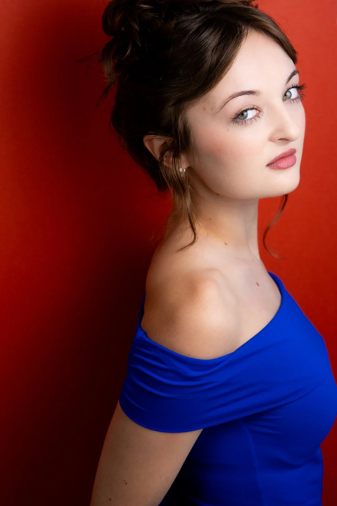 Headshot of Sarah Novak with light skin, wavy brown hair, and blue eyes, looking over her shoulder against a red background, wearing a blue off-shoulder top.