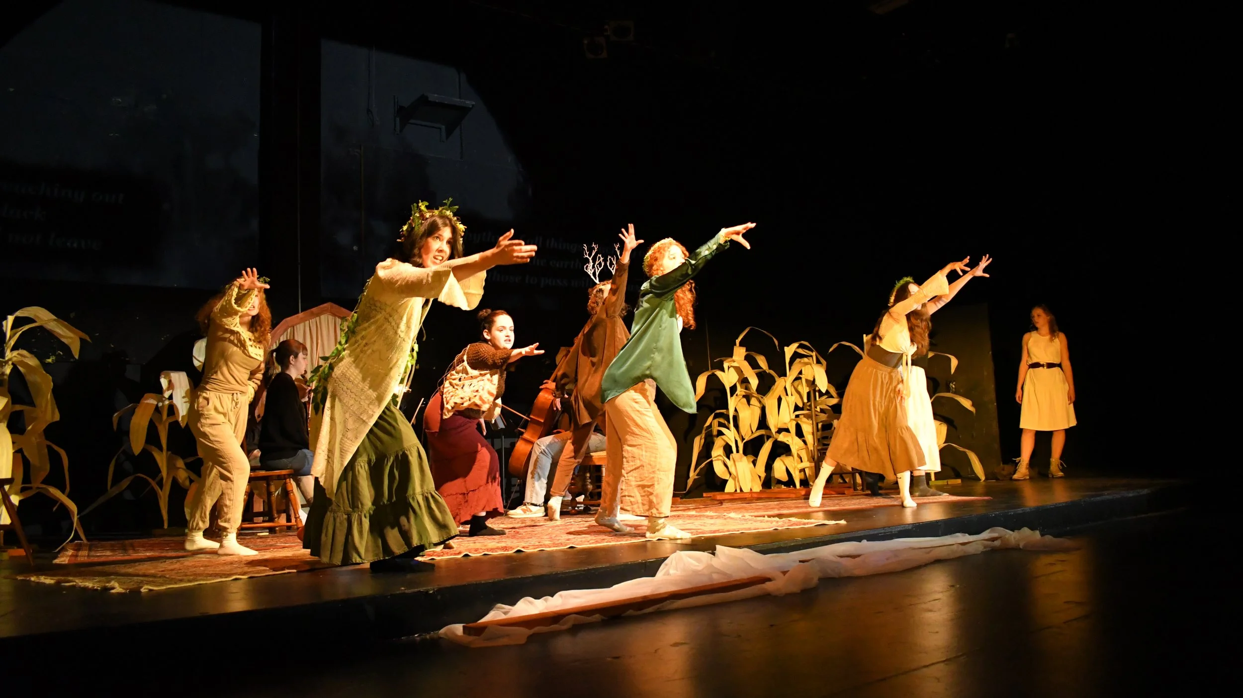 A group of children performing a play on stage with wheat decorations, dressed in costumes resembling nature and farm themes, with some seated and others standing with arms extended.
