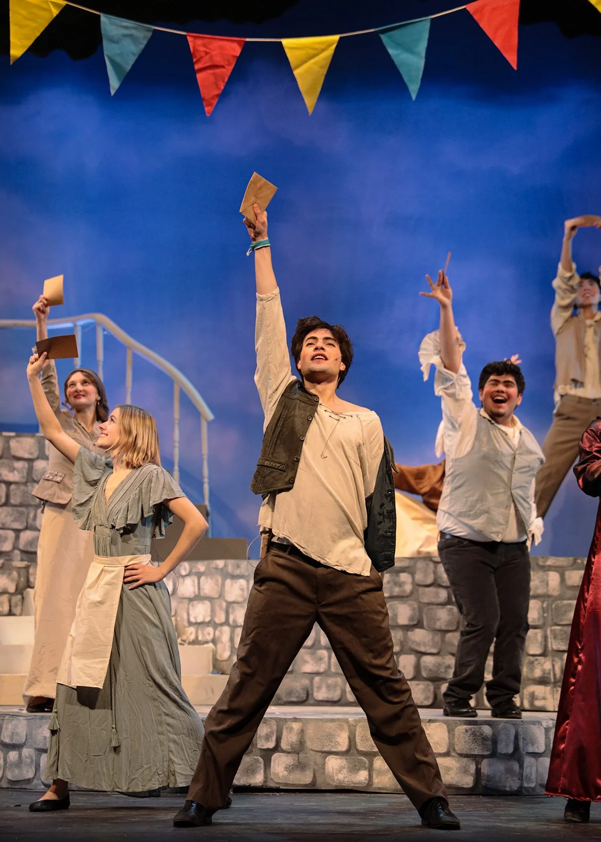 The stage scene from a theatrical performance features five actors in historical costumes, with a young man in the center raising his hand and holding a paper, on a set resembling a stone staircase and wall, with colorful pennant banners overhead and a blue backdrop.