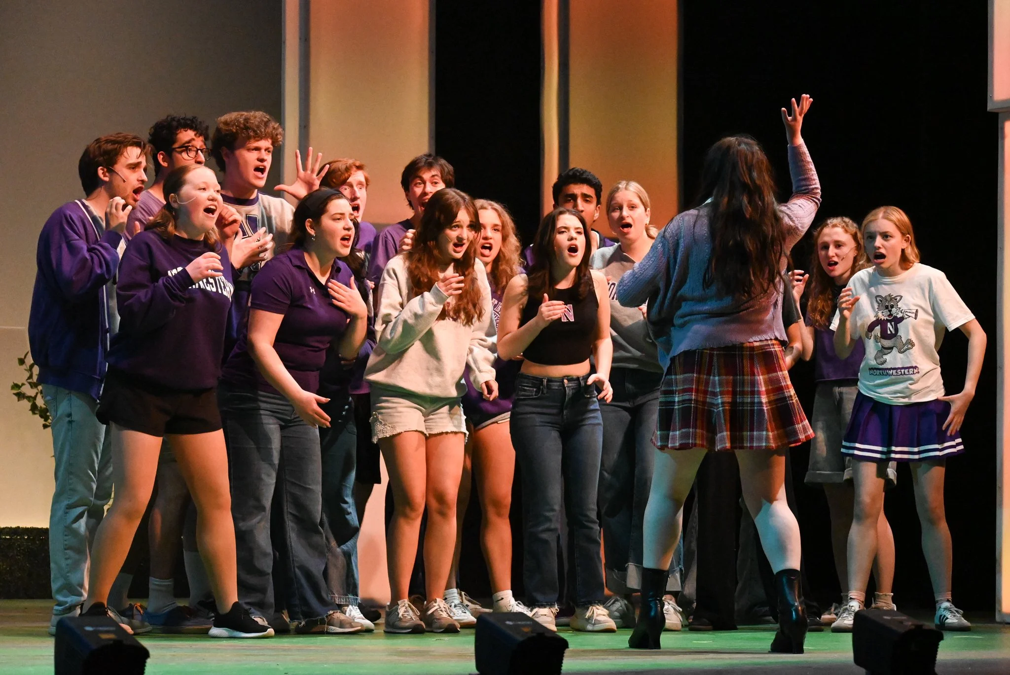 A group of young people on stage during a theater rehearsal, singing or acting with a woman in front of them, who appears to be directing or leading the group.