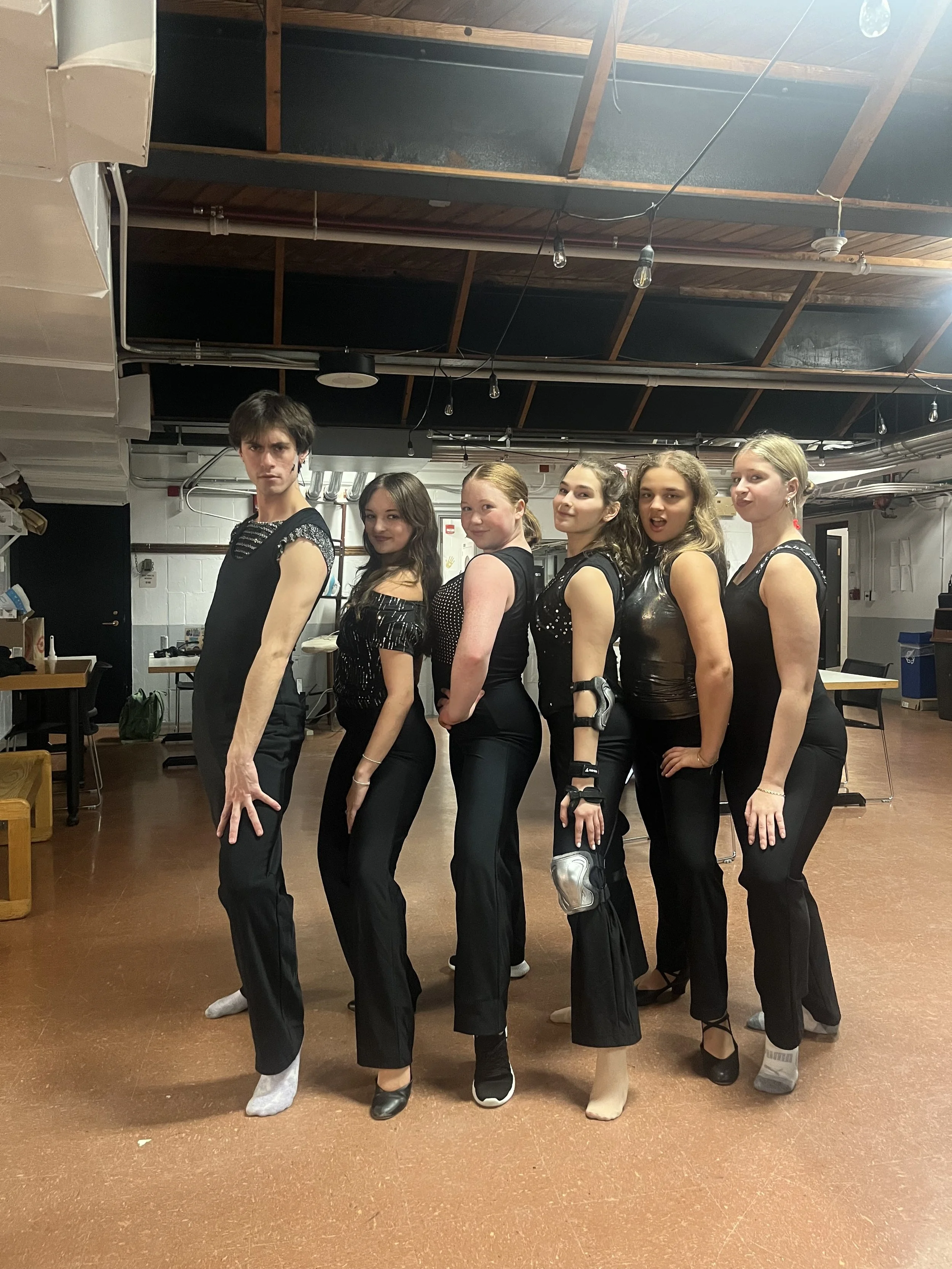 A group of six young women standing side by side in a room with a wooden ceiling, dressed in black outfits, some wearing dance or performance costumes, posing for a photo.