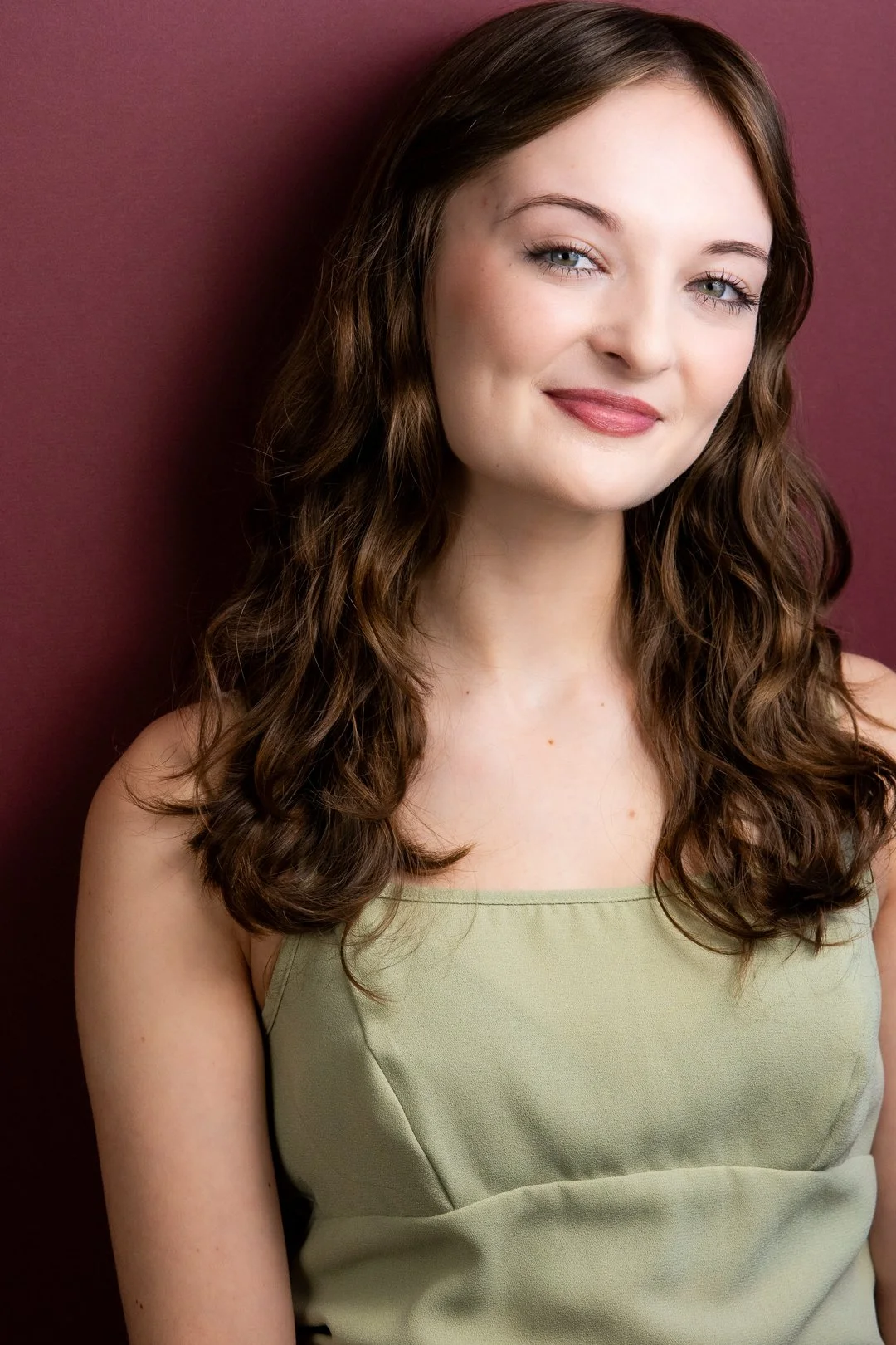 Headshot of Sarah Novak with brown curly hair, fair skin, blue eyes, wearing a light green sleeveless top, standing against a dark maroon background.