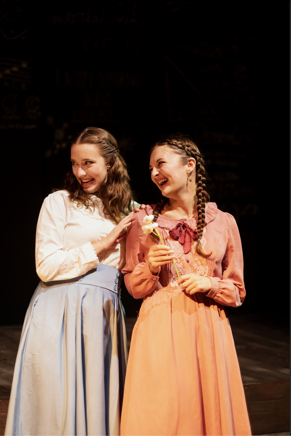 Two young women dressed in vintage-style costumes are smiling and interacting on stage, one holding flowers, with a dark backdrop and stage lighting.