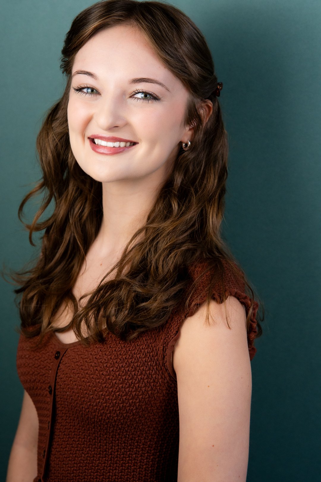 Headshot of Sarah Novak with long, wavy brown hair, blue eyes, and fair skin, smiling and wearing a rust-colored textured sleeveless top, against a dark teal background.