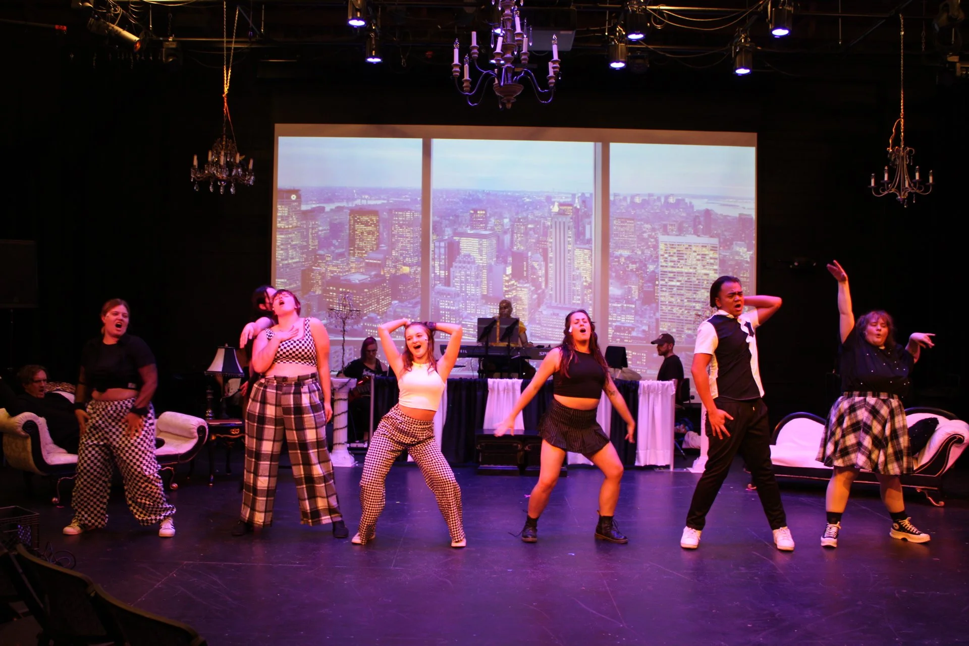 Performers on stage dancing with a city skyline backdrop, some wearing checkered pants or skirts, in a theater with curtains and chandeliers