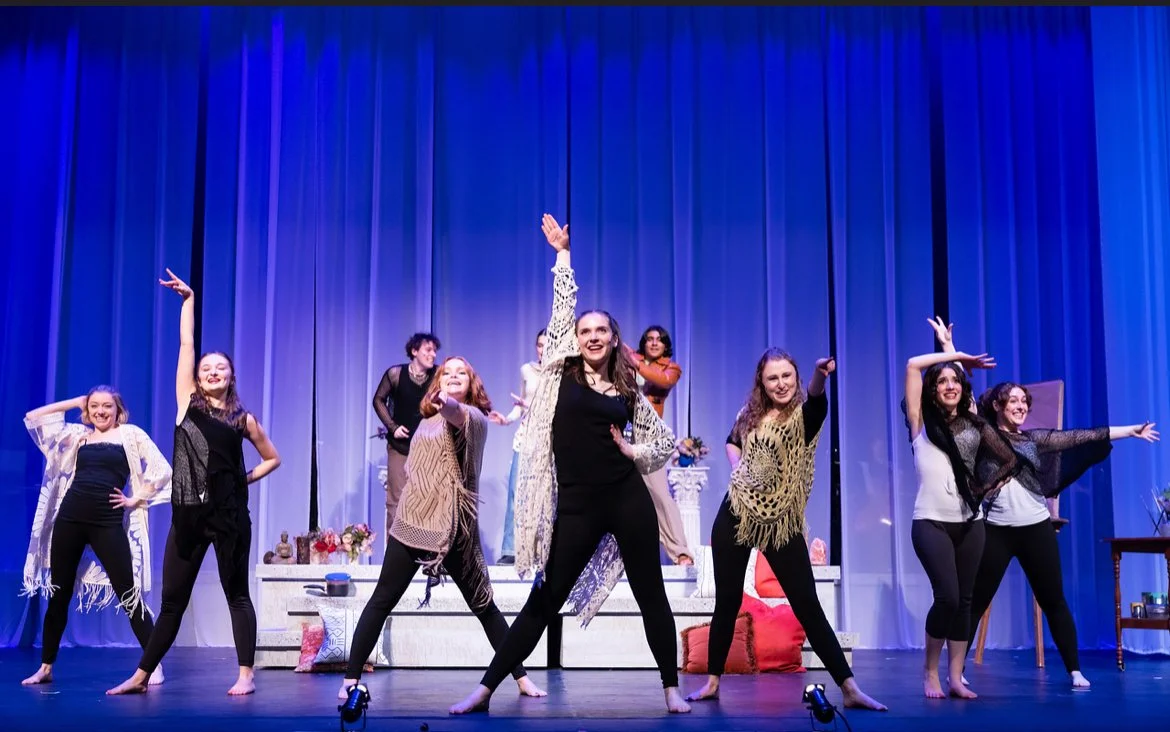 Group of women performing on stage with blue curtains in the background, various furniture, and props.