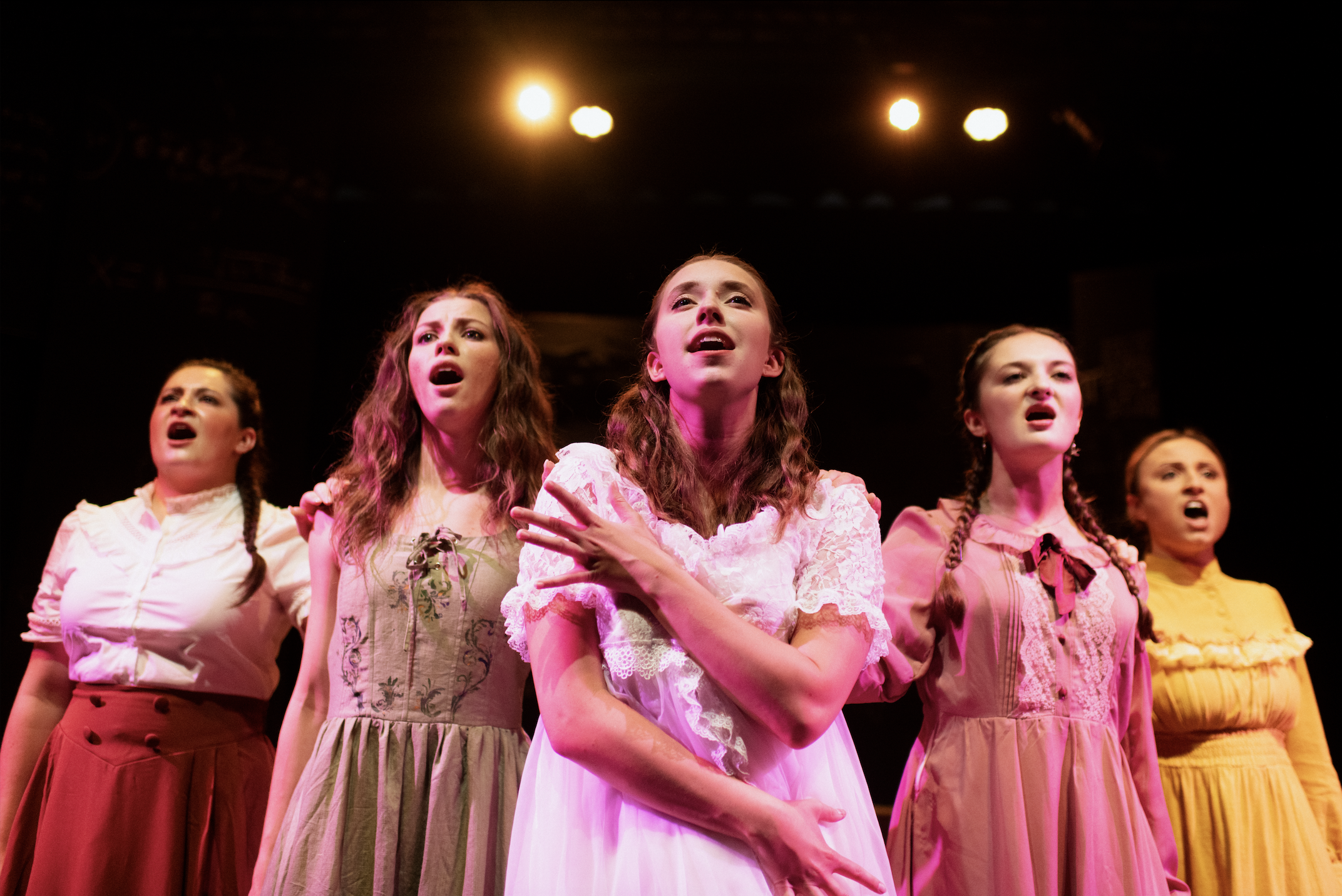 Five young women perform on stage, singing with expressive emotions. They wear vintage-style dresses in various colors, including white, green, pink, and yellow. The stage is dimly lit with warm lights in the background.