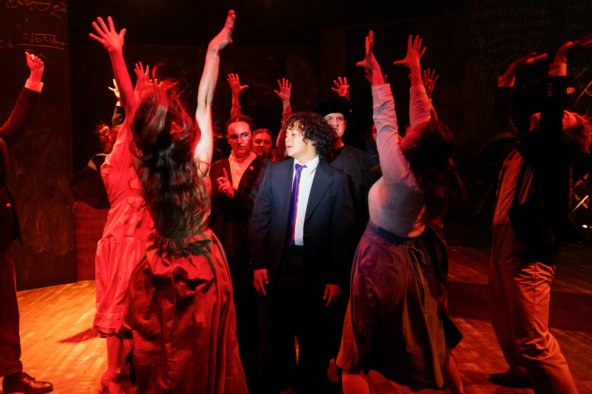 A group of people on stage participating in a drama or musical, with some raising their hands and others standing in a circle, under dramatic red lighting.
