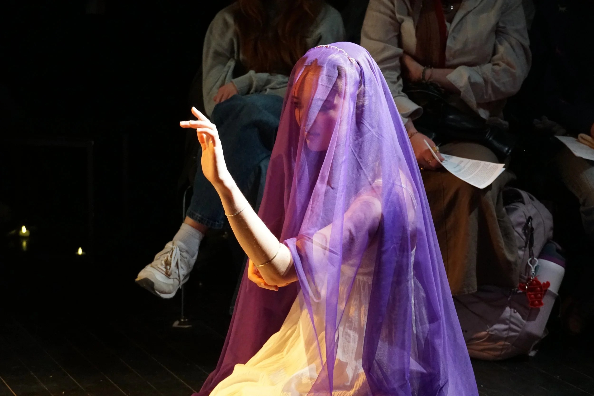 A woman dressed in a purple veil and cream dress sitting on the floor, with her eyes closed, and her hand raised gracefully. People in the background are seated, some holding papers.