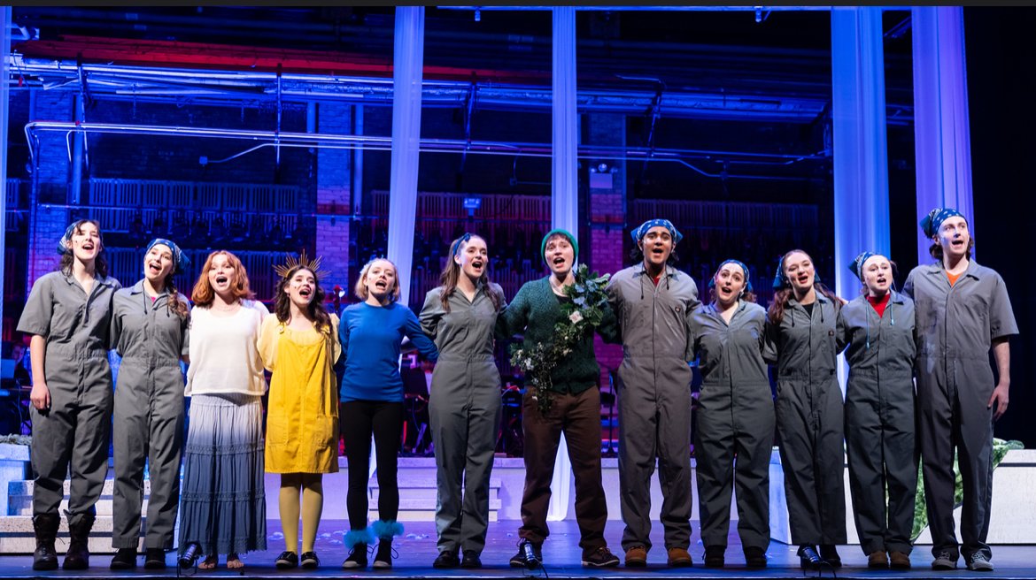 Group of performers on stage, singing with their arms around each other, in a theatrical production with colorful lighting.