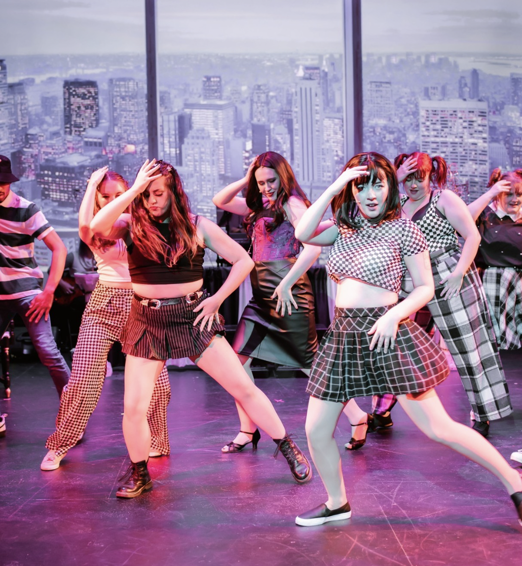 Young women performing a choreographed dance on stage with a cityscape backdrop.