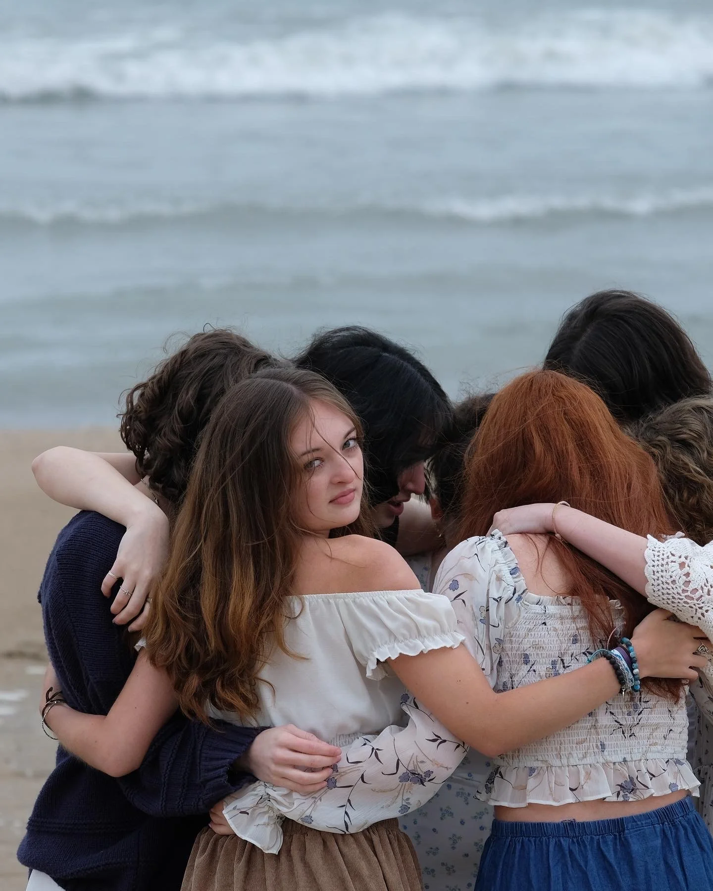 Group of young women embracing each other on the beach with the ocean in the background.