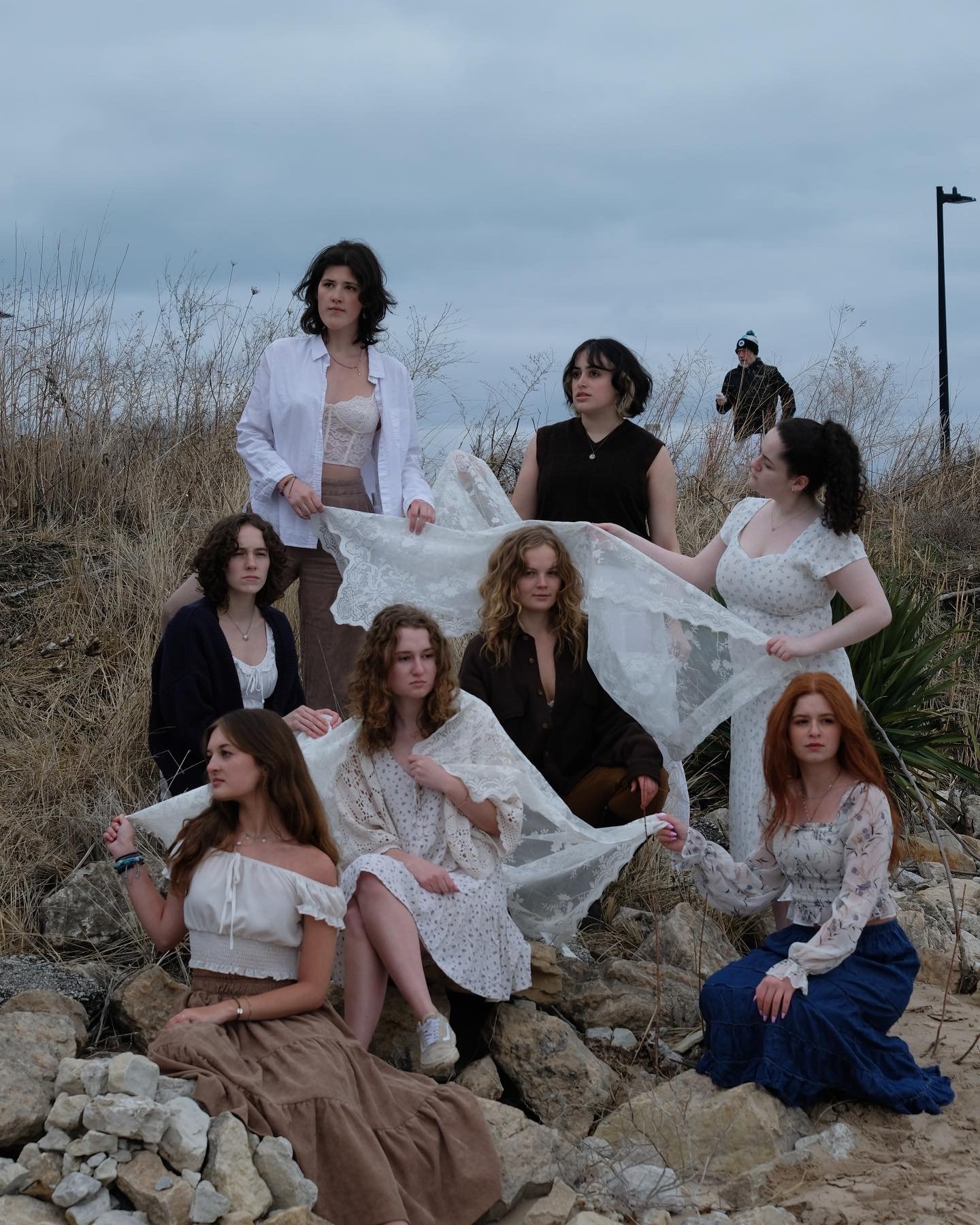 Group of nine young women outdoors on rocky terrain with dry grass and overcast sky, some holding lace fabric, posing for a photo.