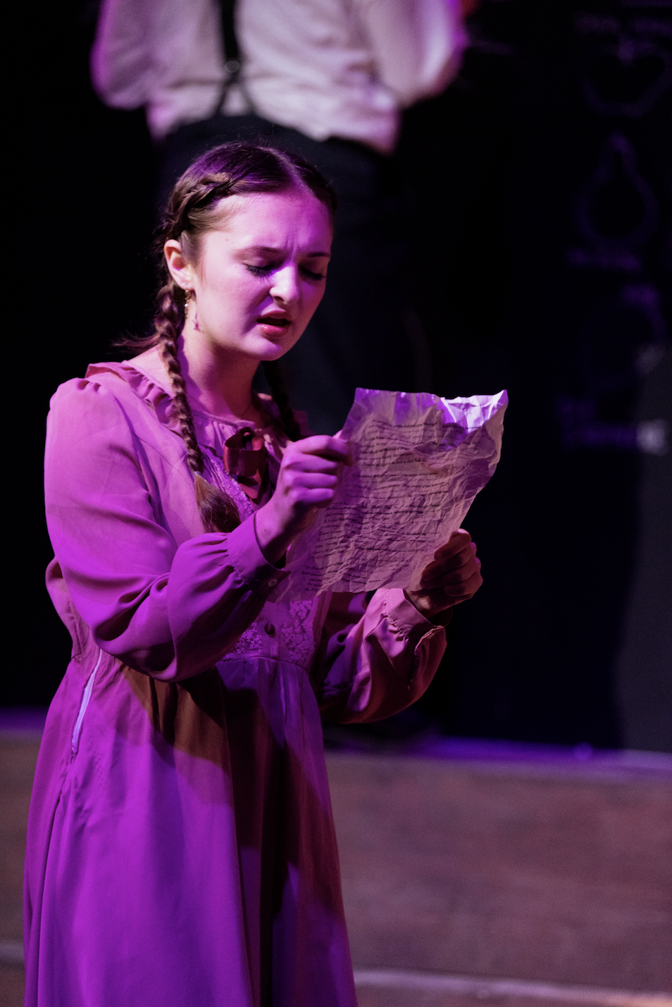 A young woman with braided hair, dressed in a pink dress, is standing on stage and reading a crumpled piece of paper with a distressed expression.