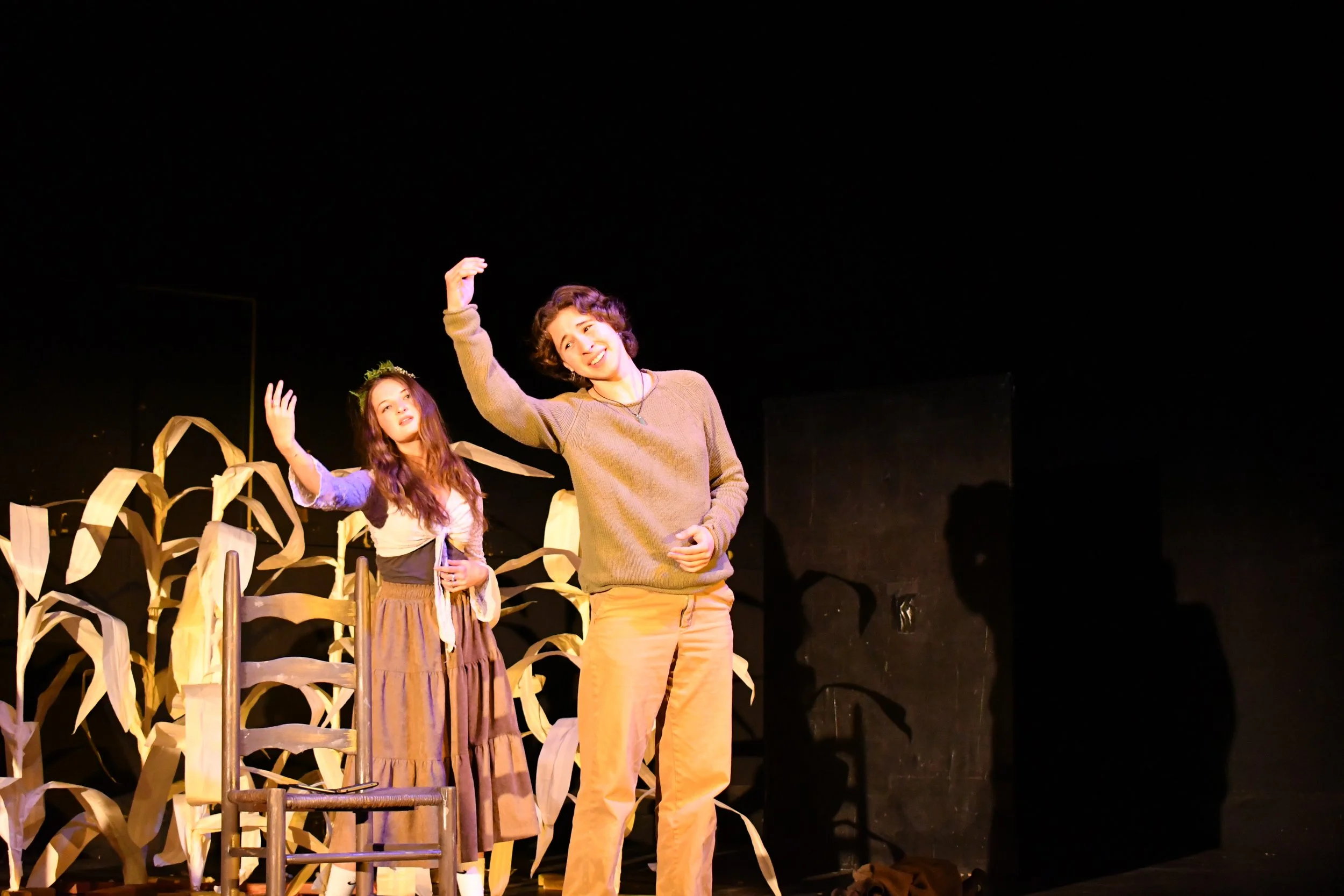 Two women on stage, one playing a character with a crown of greenery, standing among dried corn stalks, with a chair nearby, stage lighting illuminating them against a dark background.
