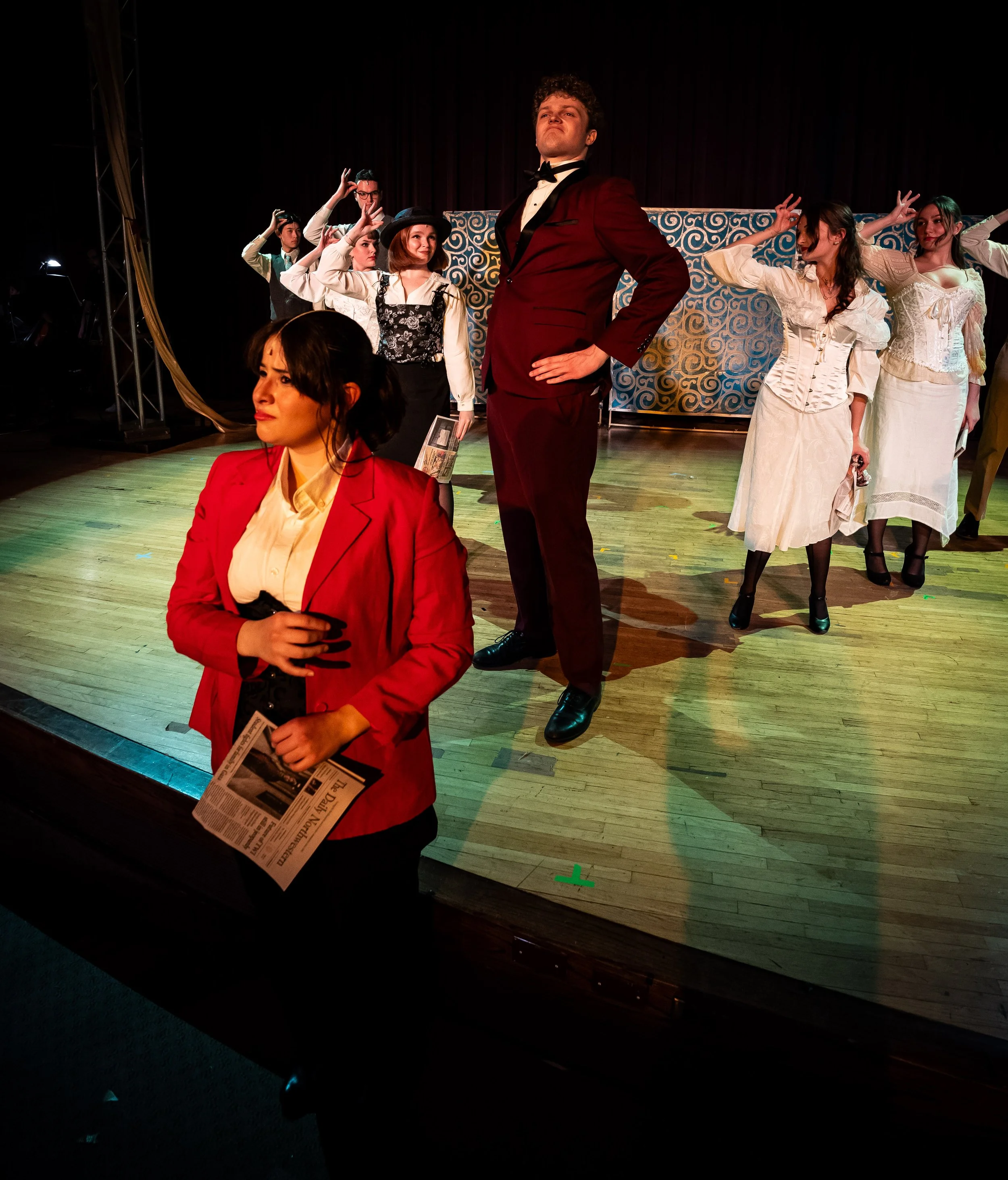 Group of people on stage in period costumes, with one woman in a red blazer in the foreground, and others standing or sitting behind her, performing in a theatrical production.