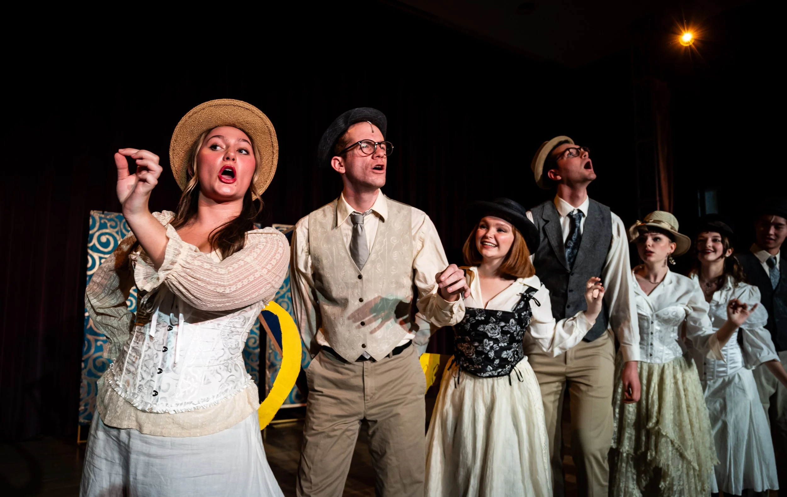 The cast of a theatrical play dressed in vintage costumes, performing on stage with expressive facial gestures.