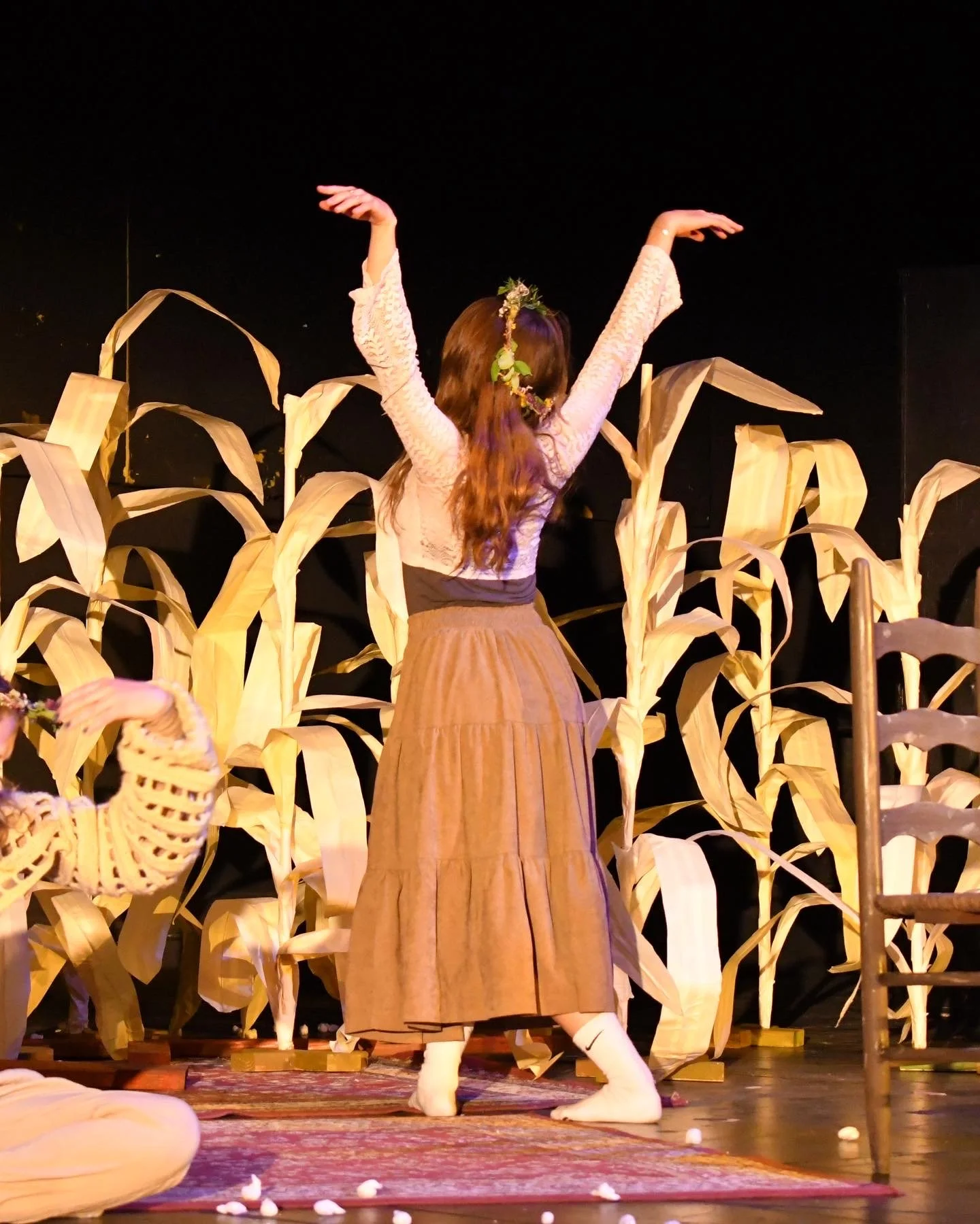 Sarah Novak in Creating Small Thunder by Mya Vandegrift at Northwestern University. A woman in a brown skirt and white top dancing on stage with arms raised, surrounded by wooden corn stalk decorations, with a black background.