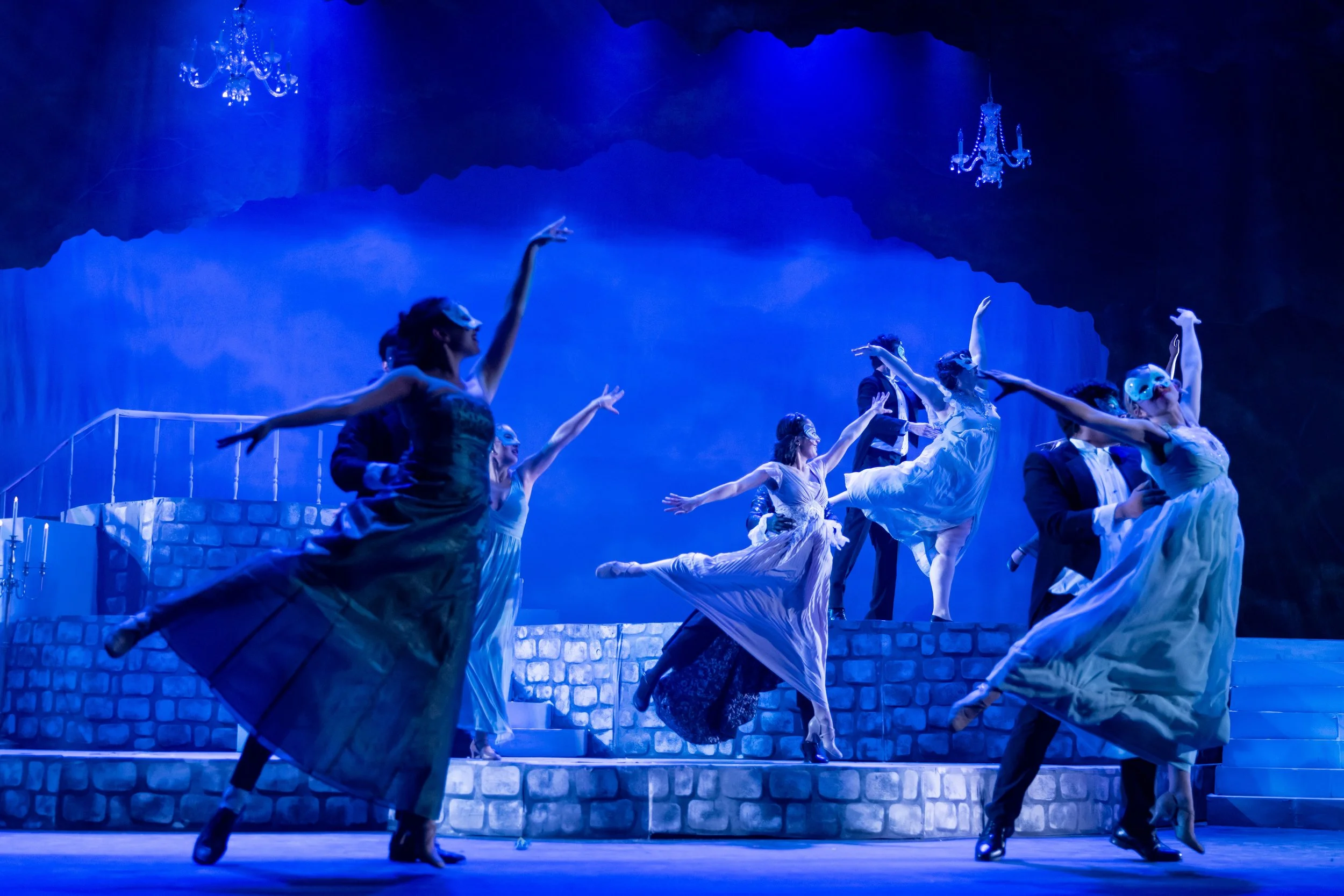 Ballet dancers performing on stage with blue lighting, stone steps, and chandeliers hanging from the ceiling.