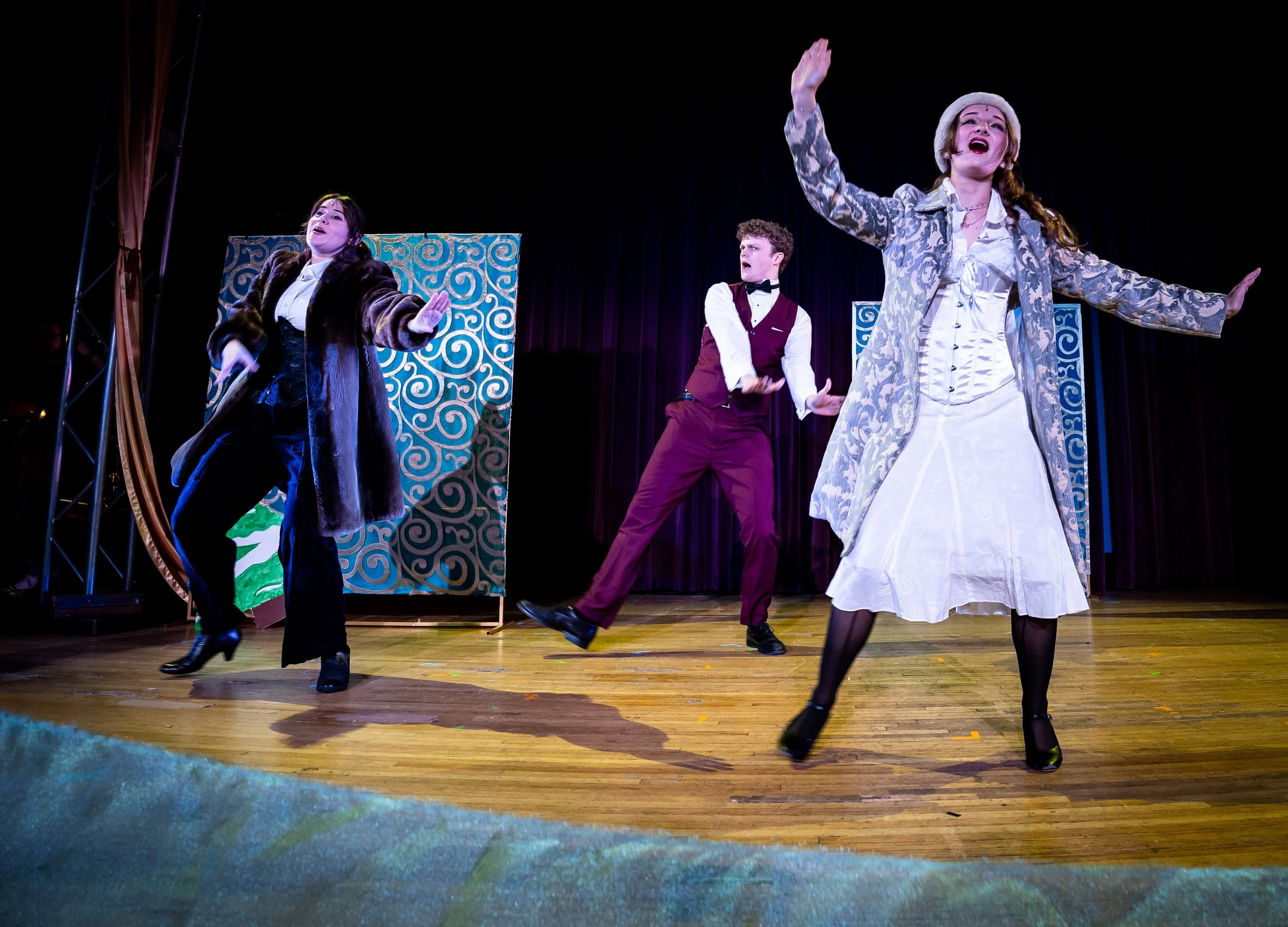 Three performers on stage engaged in a theatrical dance or performance, with two women in costume and a man in a vest and bow tie, against a backdrop with swirling designs.