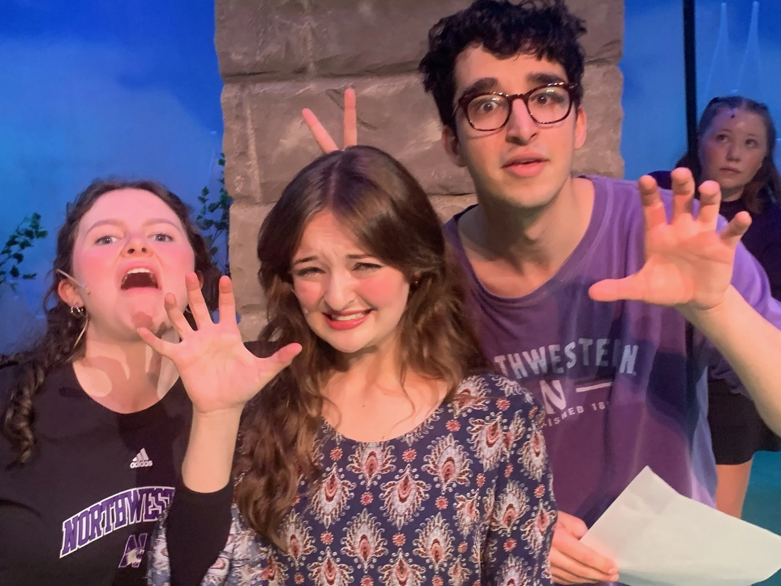 Three young people, two women and one man, making funny faces and gestures towards the camera in an indoor setting with blue lighting and a stone wall backdrop. The woman on the left has long braided hair, the woman in the middle has wavy brown hair, and the man on the right has curly dark hair and glasses.