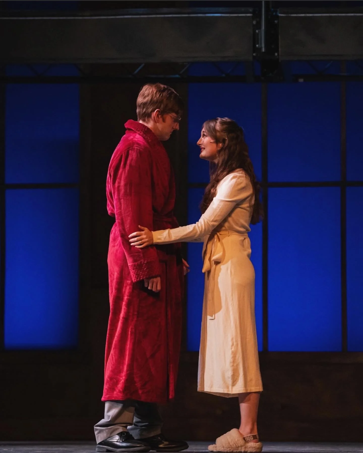 Sarah Novak and Garraway Nance standing close together on stage, smiling at each other. The man is wearing a red robe and glasses, while the woman is in a beige dress and slippers. The background features blue window panels.
