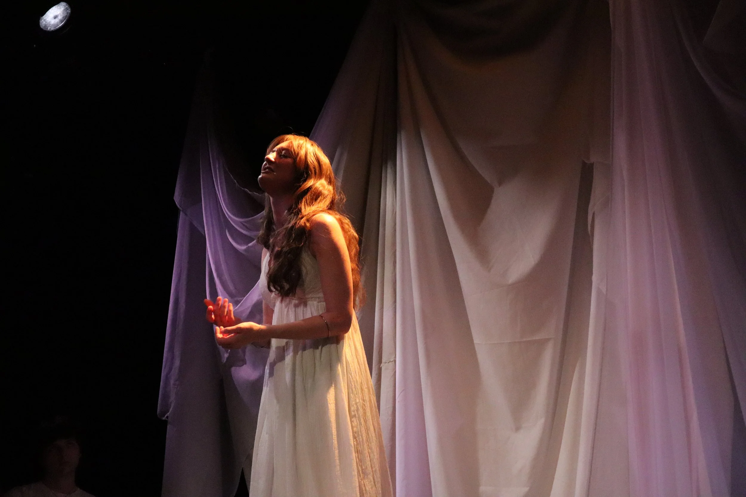 A woman with long, wavy brown hair stands on stage, wearing a white dress, illuminated by a spotlight, with large draped fabric backdrop behind her.