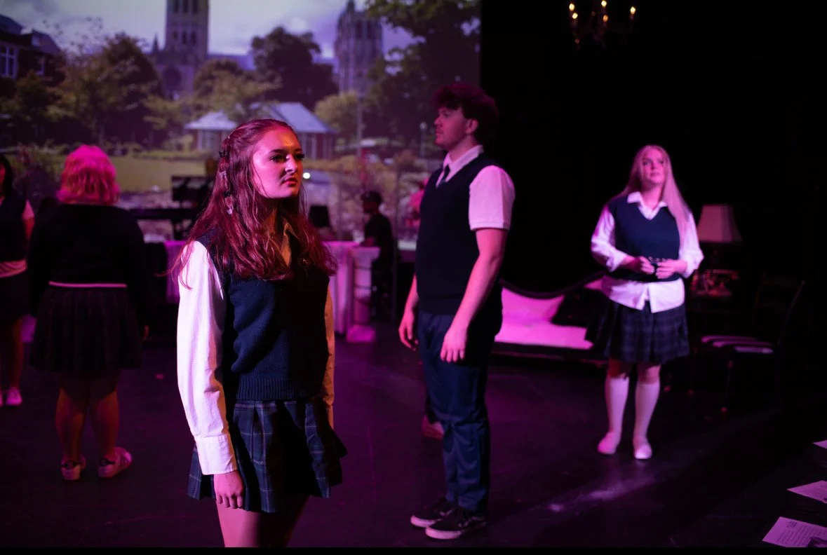 Students in school uniforms on stage during a play, with a backdrop of a town scene projected behind them.